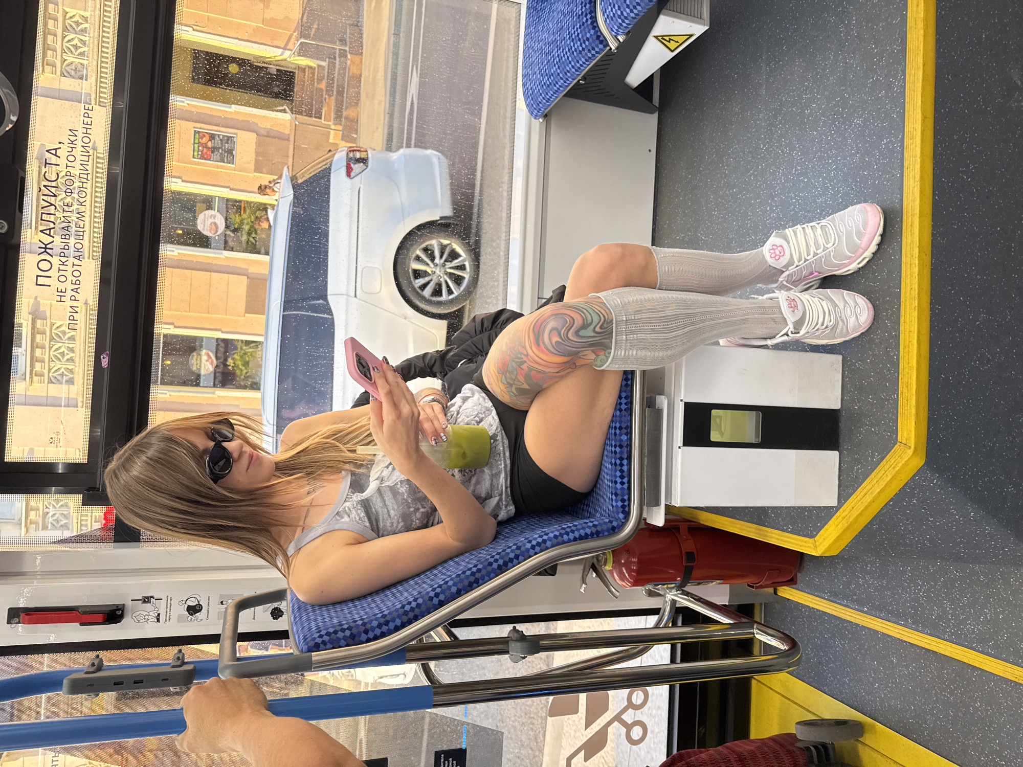 A young woman is seated on a blue and checkered bus seat, looking down at her pink smartphone. She wears black shorts, a grey tank top with a graphic print, sunglasses, and white socks that reach just below her knees. Her legs are adorned with colorful tattoos, and she is holding a green beverage in a clear plastic cup. In front of her is a fire extinguisher. The bus interior features yellow safety markings on the floor and blue seats. Outside the window, a white minivan drives past a building with architectural details and signage in Russian. The scene takes place in Moscow, Russia.