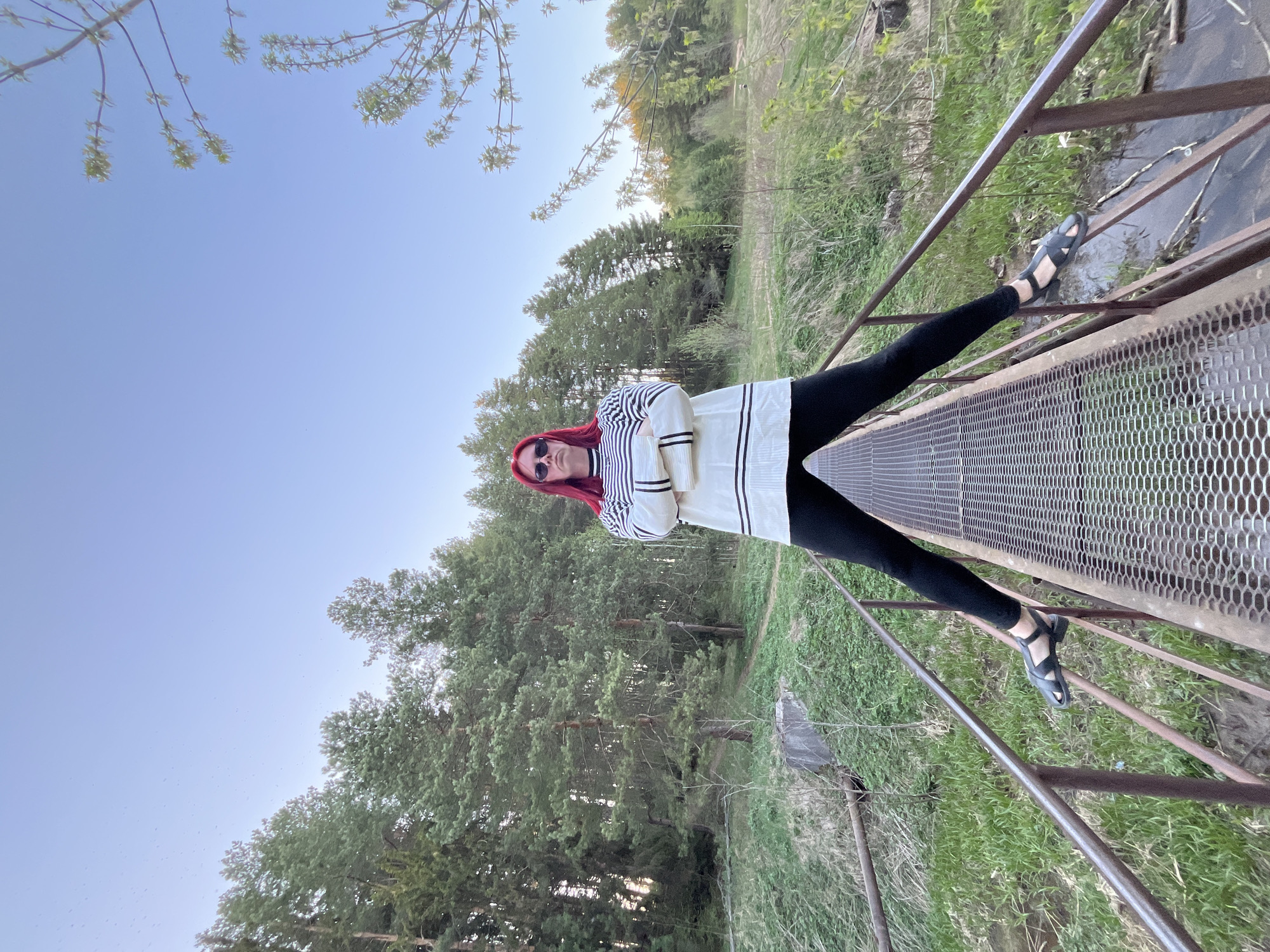 A woman with red hair and sunglasses stands on a metal bridge with her legs spread wide, extending to the railings. She wears a striped top and black leggings with sandals. The bridge spans a grassy area with trees. The image was likely taken in the city of Chaikovsky, Russia. The sky is light blue, indicating daylight.