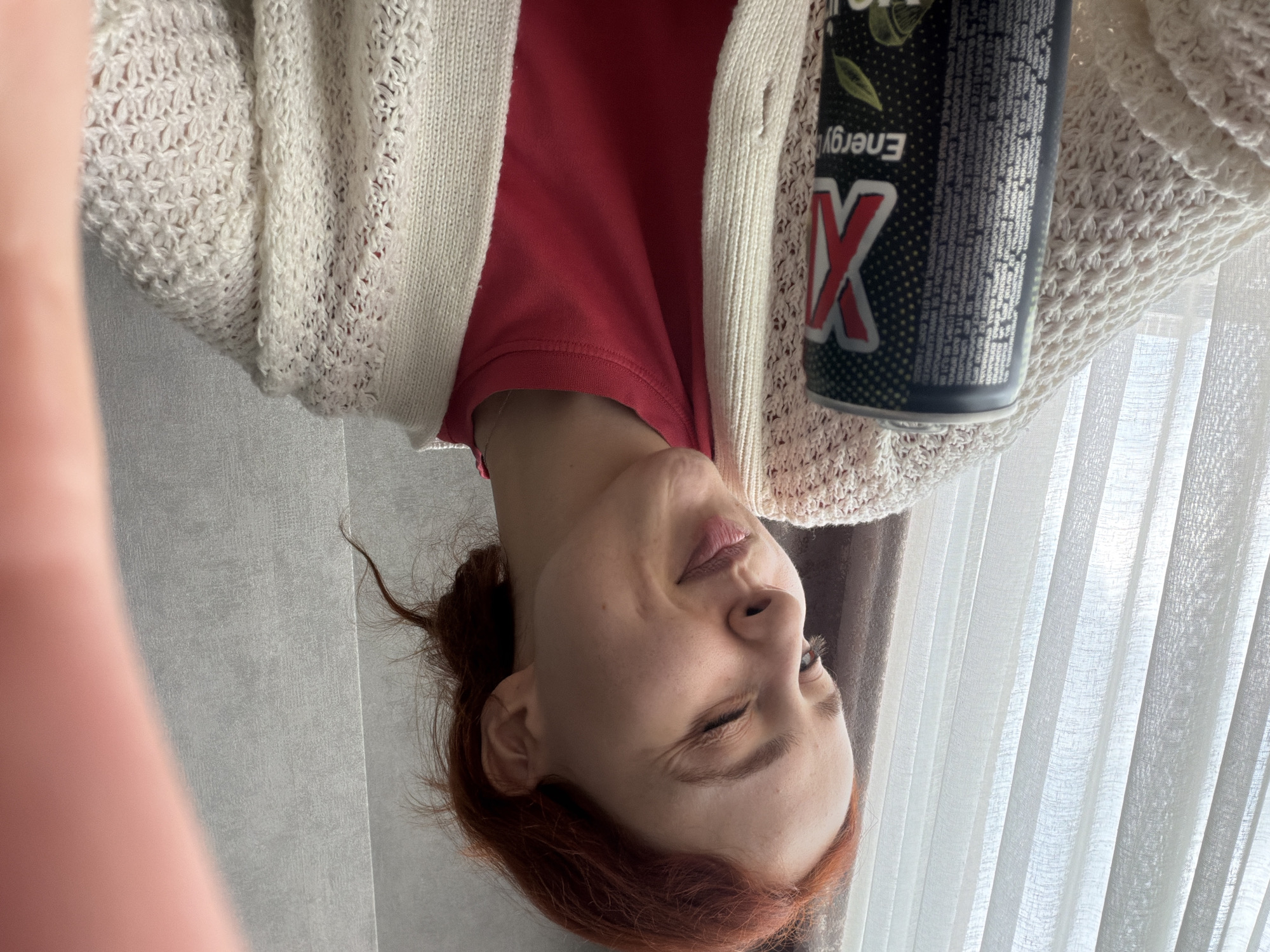 A woman with red hair, wearing a red shirt and a white knit cardigan, is holding a can of "X Energy" drink. She is indoors, likely near a window with white curtains. The location may be in Tbilisi, Georgia.