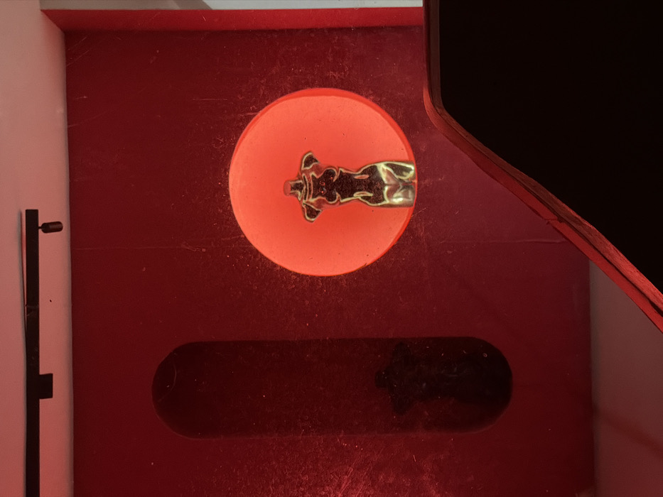 This image captures an interior scene, likely a gallery or retail display, located in Kemenuh, Indonesia. The dominant feature is a dark red wall. A prominent metallic, golden-hued, headless and limbless female torso figurine is positioned within a circular cutout on this wall, strongly backlit by an intense orange-red light. To the left, a dark, elongated, arched recess contains a dimly visible, dark silhouette of a similar female torso figurine. Overhead, a black track light fixture with a single spotlight head is mounted on a white ceiling. In the lower right foreground, an angular, dark surface with a reddish-illuminated edge is partially visible, suggesting a counter or display plinth. The wall surface exhibits minor dust or texture.