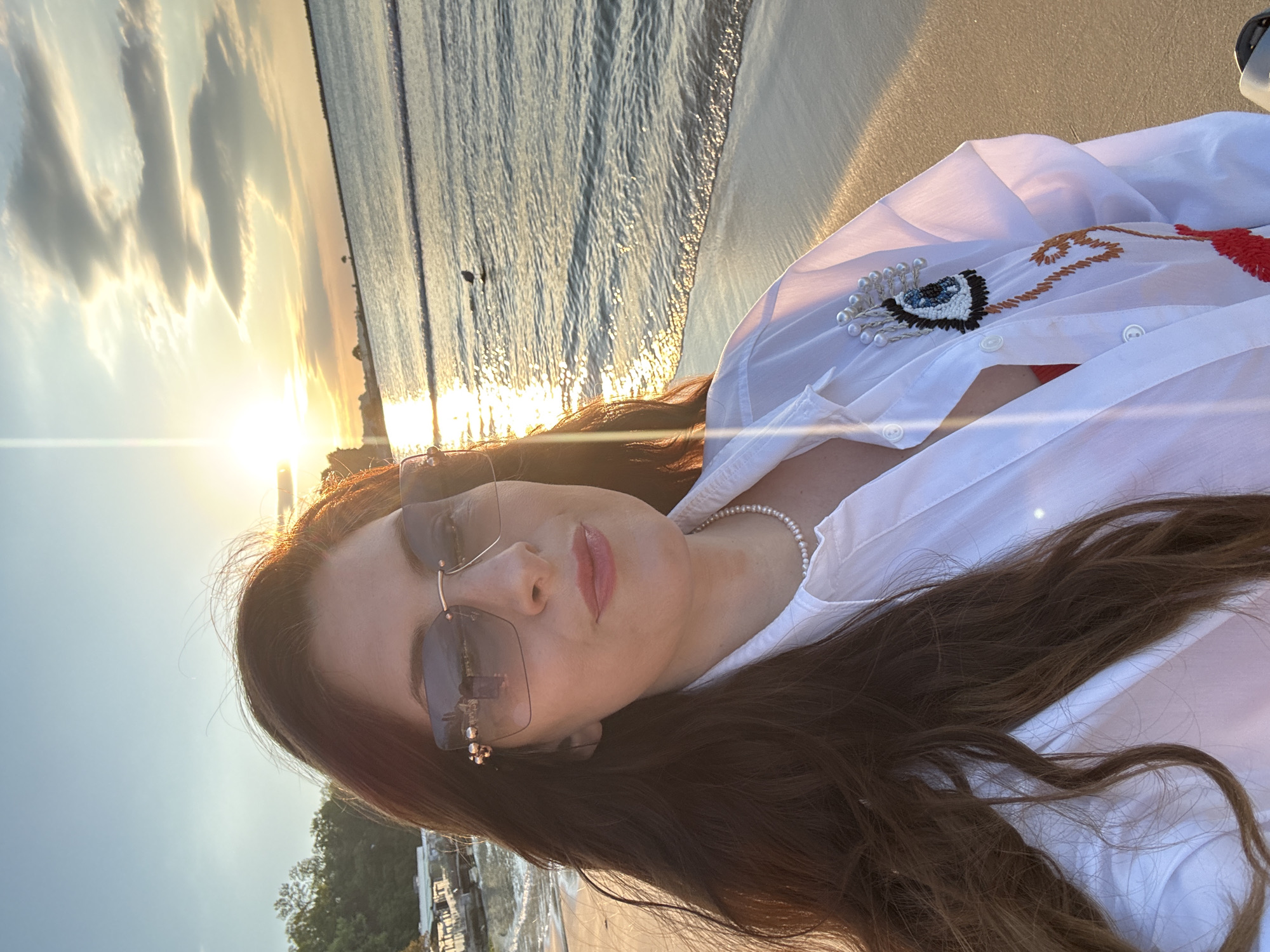 A woman with long brown hair is wearing sunglasses and a white shirt, posing on a beach at sunset. The sun is setting over the sea in the background. The sky is a mix of orange, yellow, and blue. The woman is wearing a pearl necklace and earrings. Her shirt has an embroidered design on the front. The location appears to be a beach in Varna, Bulgaria.