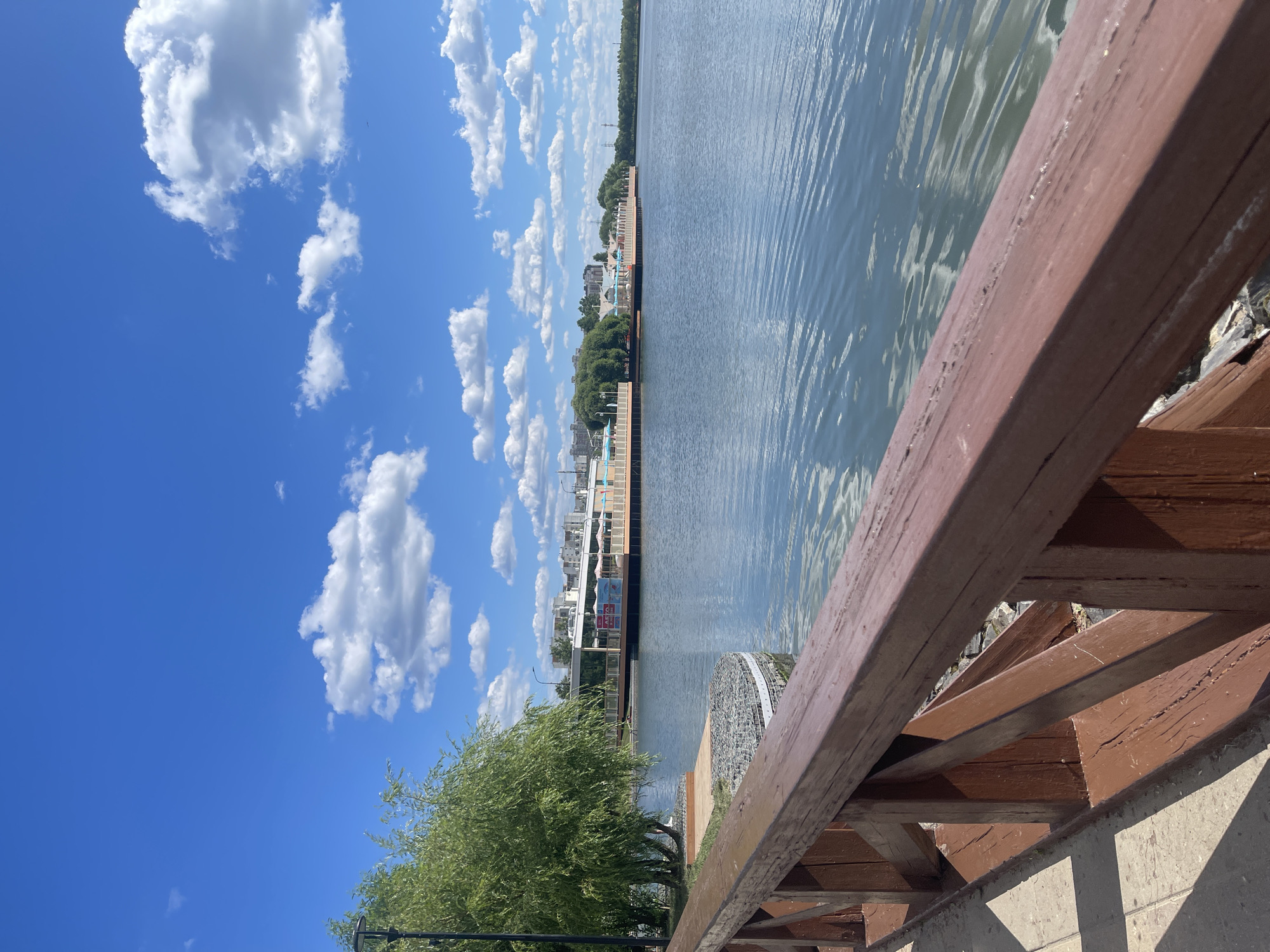 The image depicts a sunny day with a blue sky filled with white clouds. In the foreground, a wooden railing, painted brown, runs diagonally across the frame. Beyond the railing is a paved walkway and a body of water, likely a lake or river, with ripples reflecting the sky. Across the water, a long wooden pier or boardwalk extends, lined with buildings that appear to be restaurants or cafes with white awnings. Trees are visible behind the buildings, and in the distance, several apartment buildings suggest an urban setting. The scene is tranquil and appears to be a public park or recreational area. This location is likely in Moscow, Russia, given the architectural style of the buildings and the presence of such waterfront recreational spaces. There are no people visible in the image.