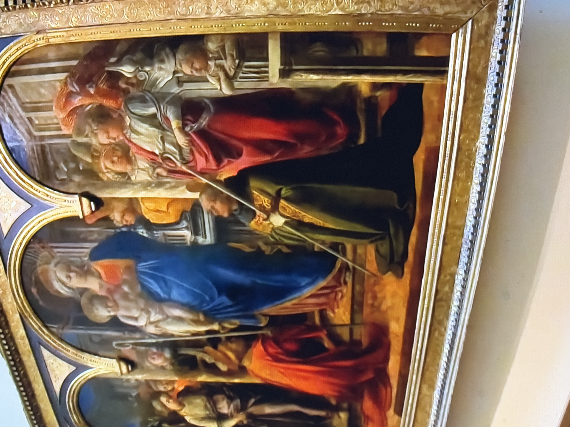 A painting depicts a religious scene within a gold-ornamented, arched frame. In the central arch, the Virgin Mary, wearing a blue robe and an orange garment, cradles the infant Jesus. To Mary's right, a figure in a red robe holds a staff. To the left of Mary, another figure kneels in a red robe, holding a staff. Behind these figures, other individuals are visible, some with yellow garments. To the far right, a young child stands by a column, leaning on a railing. The painting's background features architectural elements and a warm, orange-toned floor. The scene is set indoors, likely a church or a formal hall.