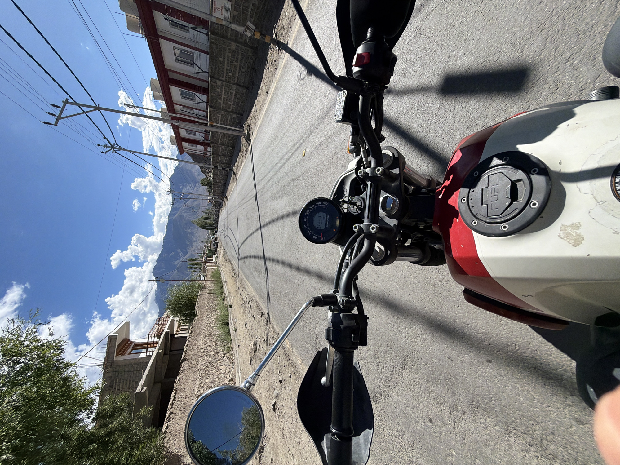 The image captures a first-person perspective from a motorcycle on a paved road in Leh, India. The road appears to be a main thoroughfare, with buildings lining both sides. On the left, there's a stone wall and a multi-story building under construction. On the right, a long, white building with red trim and windows is visible, with power lines and poles running along the street. In the background, majestic mountains form a dramatic backdrop under a bright blue sky with scattered white clouds. The motorcycle's handlebars, instrument cluster, and fuel tank are in the foreground, indicating movement. The overall scene depicts a sunny day in Leh, with typical urban and mountainous landscape elements.