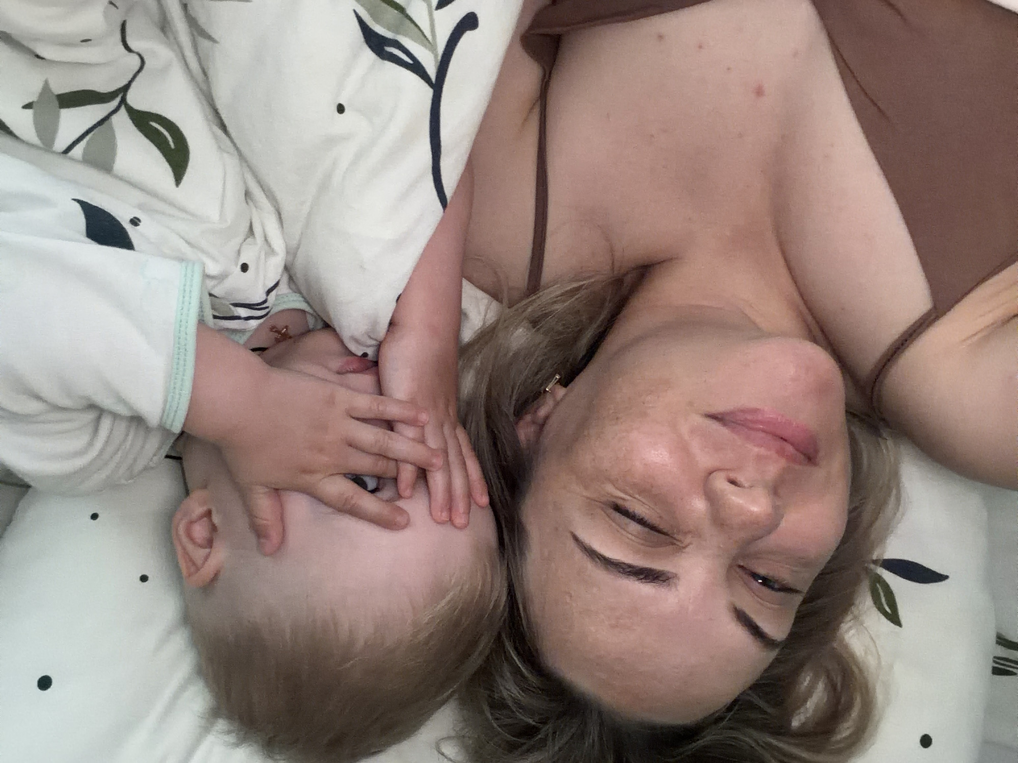 An intimate overhead photograph captures a woman and a young child lying closely together on a bed, bathed in soft, natural light, suggesting a tranquil moment indoors in Votkinsk, Russia. The adult woman, positioned on the right side of the frame, has fair skin with visible freckles and light brown hair. She wears a simple brown or dark beige camisole. Her eyes are partially closed in a gentle, contented smile, conveying warmth and relaxation.
To her left, a young child with light blonde hair is nestled against her. The child is dressed in a white long-sleeved top with a light green trim on the cuff. Playfully, or perhaps shielding from light, the child covers their eyes with both hands. The bedding is white, adorned with a stylized pattern of dark green and gray leaf motifs and scattered small black dots. The soft illumination suggests either early morning or late afternoon, or diffused daylight within the room. The image emanates feelings of affection, tenderness, and a close familial bond. No discernible text is visible within the frame.