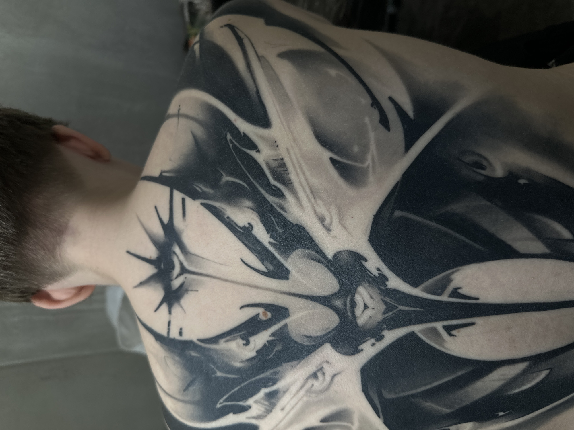 A close-up shot shows a person's back and the back of their neck, which features a large, black and gray tattoo. The tattoo is abstract, with sharp lines and gradients creating a dynamic design. The person's hair is short and light-colored. The background is out of focus and indistinct, appearing to be a neutral gray surface. The location is not discernible from the image.
