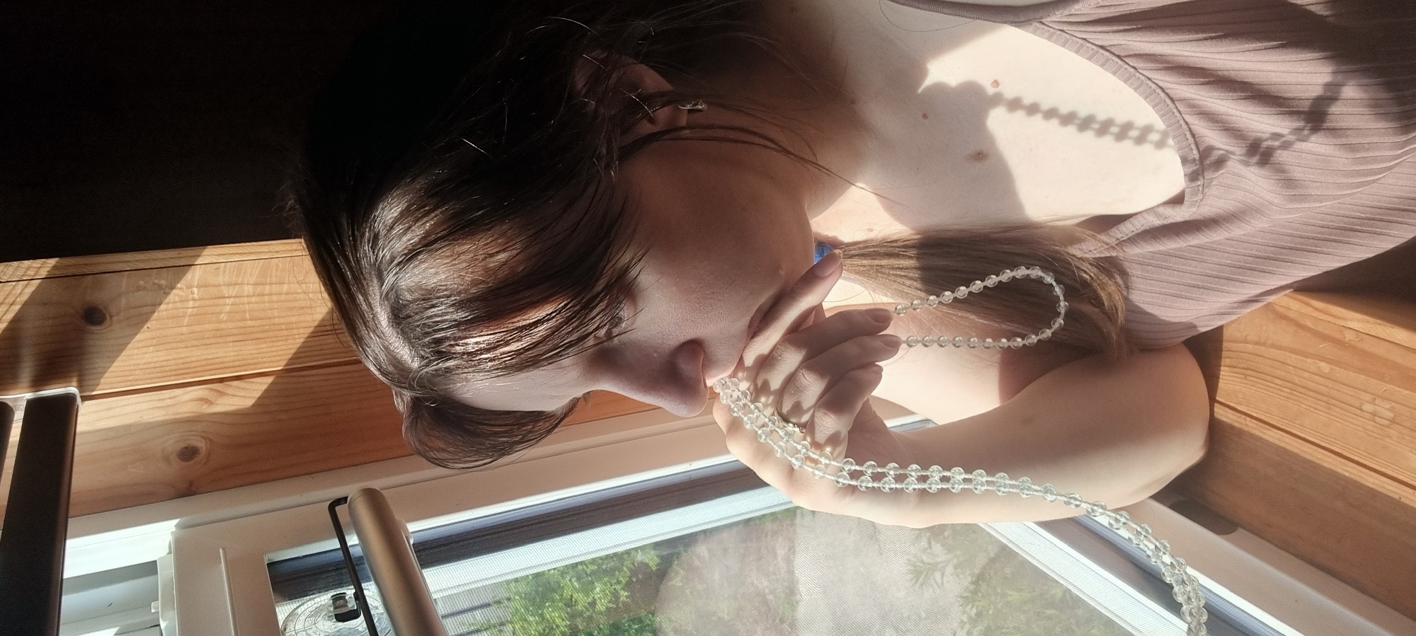 The image, rotated 90 degrees clockwise, captures a close-up of a person in an indoor setting bathed in sunlight. The subject, seen from the upper chest, has long dark brown hair and fair skin. They are wearing a light brown ribbed tank top or camisole, and an earring is visible in their left ear, which also appears to have a small blue earbud or similar device.
The person's left hand holds a long strand of clear, faceted beads, possibly a necklace or rosary, which also drapes around their neck and chest. A ring is visible on their left ring finger. Their head is tilted downwards, suggesting a contemplative or focused gaze towards the beads or out the window.
The background features a wooden wall or paneling, indicative of an interior space. Below the person, a window with a screen is visible, through which green foliage can be seen, hinting at an outdoor natural environment. Two metallic, cylindrical objects stand near the window, possibly parts of a fixture like a lamp or window shade.
Bright, direct sunlight streams in, creating strong, defined shadows of the beads on the person's skin and a sharp, rectangular shadow on the wooden wall. This indicates it is daytime, likely morning or afternoon. The scene evokes a sense of peace and quiet introspection in a sunlit room in Dunnae-myeon, South Korea. There is no visible text.