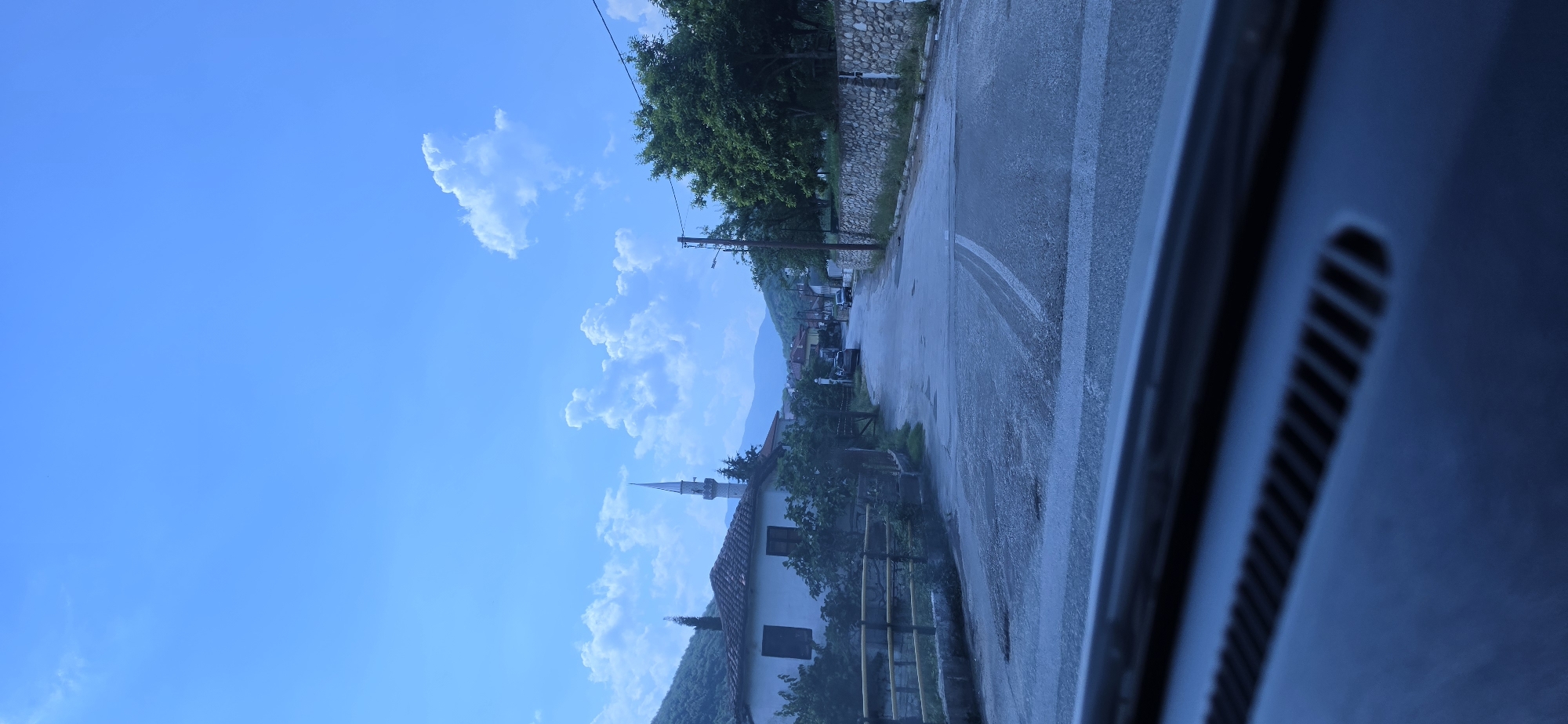 The image depicts a rural street scene in Melivoia, Greece, captured from the interior of a vehicle, evidenced by a dark dashboard with vents at the bottom of the frame. A paved road, appearing worn with some visible patches, stretches into the distance, with a single white road marking partially visible in the center.
On the left side of the road, a traditional white building with a terracotta tiled roof and dark-framed windows is prominent. Behind this building, a slender, light-colored minaret with a pointed spire rises into the sky, indicating the presence of a mosque. Green trees and shrubbery are visible around the building and along a wooden fence lining the road. Steep, tree-covered mountains form the immediate backdrop on the left.
On the right side of the road, a dry-stone wall extends, topped with additional fencing, with lush green trees behind it. A utility pole with electrical wires stands near the wall. In the distance, other buildings and further mountains are discernible. The sky is bright blue with scattered white, cumulus clouds. No individuals are visible in the scene.