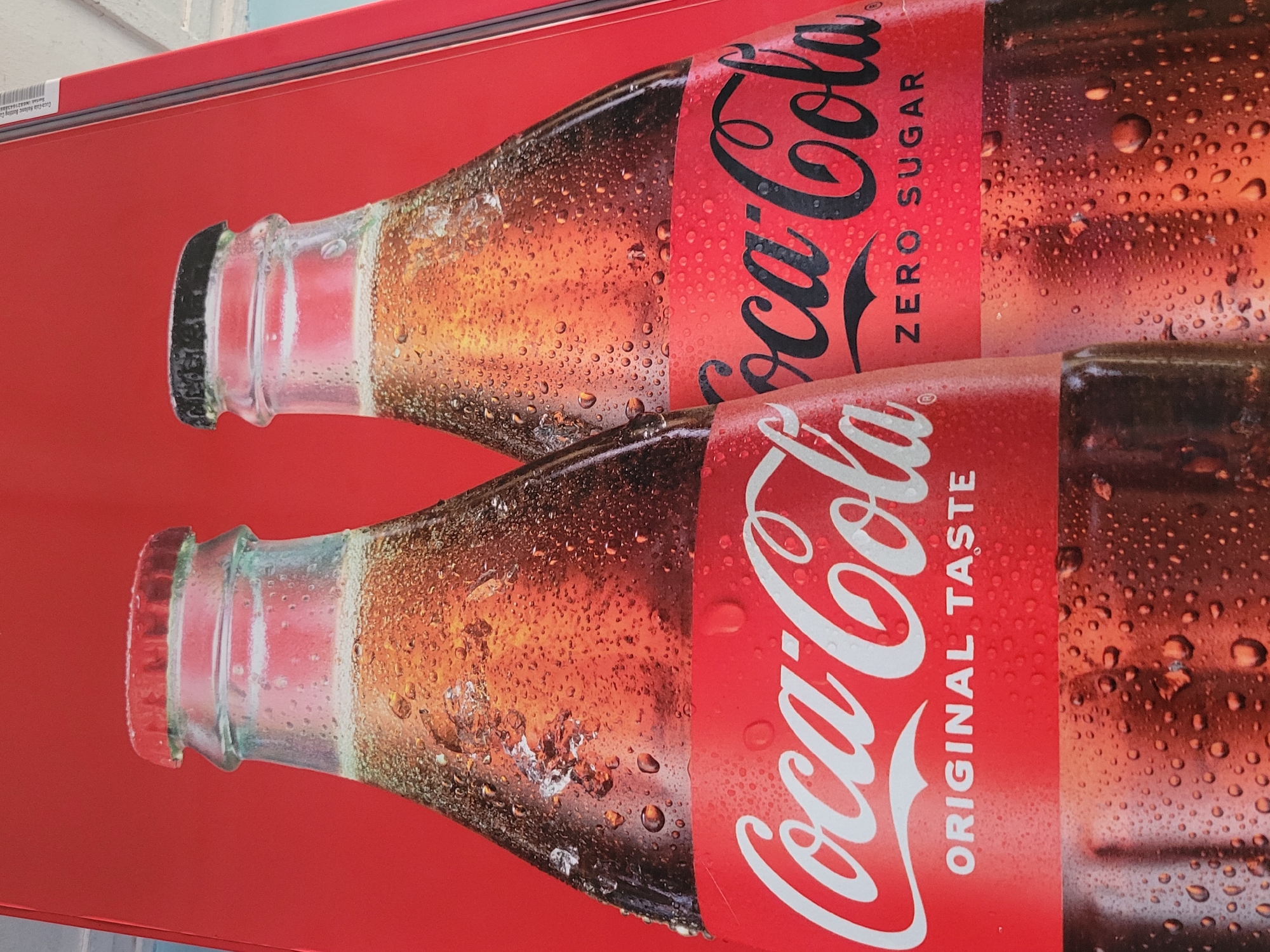 This media file features a close-up, eye-level shot of two Coca-Cola bottles against a bright red background. The bottles are depicted as if they are sweating, with numerous water droplets adorning their glass surfaces and labels, suggesting they are chilled. Both bottles have their iconic red labels. The top bottle's label is partially obscured by the bottom bottle. The visible text on the top bottle reads "Coca-Cola" in large white script and "ZERO SUGAR" vertically. The bottom bottle's label also displays the "Coca-Cola" script in white, with "ORIGINAL TASTE" written vertically below it. The image is likely a promotional graphic, possibly for an advertisement or display, emphasizing the refreshing quality of the beverage. There are some faint, almost illegible text details on a white sticker in the upper left corner, potentially indicating manufacturing or serial information, with "Jos, Nigeria" visible in the OCR. The lighting is bright and even, highlighting the condensation on the bottles. There are no people or specific environmental cues visible, making the focus entirely on the product.