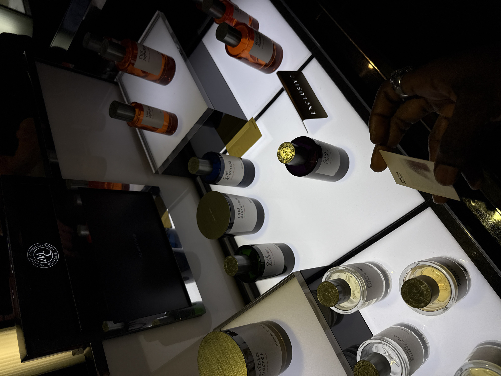 This image captures a luxury fragrance display for Maison Crivelli Parfums, located within a high-end department store, specifically identified as Harrods in Knightsbridge, London, United Kingdom.
The scene features multiple Maison Crivelli perfume bottles arranged on individual, brightly illuminated white platforms. Visible fragrances include "Safran Secret," "Oud Stallion," "Oud Maracuja," "Oud Charnel," "Rose Saltifolia," "Fleur Diamant," "Cuir Infini," and "Hibiscus Mahajád." A large black display box with the "Maison Crivelli Parfums Paris" logo is positioned at the back of the display. A small sign with the word "EXCLUSIVE" is also present on one of the platforms.
In the lower right quadrant of the image, a dark-skinned right hand, wearing a silver ring on the ring finger, holds a white paper fragrance testing strip. The strip displays a reddish stain and partially legible text, "Safran Secret," indicating a fragrance sampling action. The overall environment is dimly lit, with strategic illumination highlighting the product displays, typical of luxury retail settings.