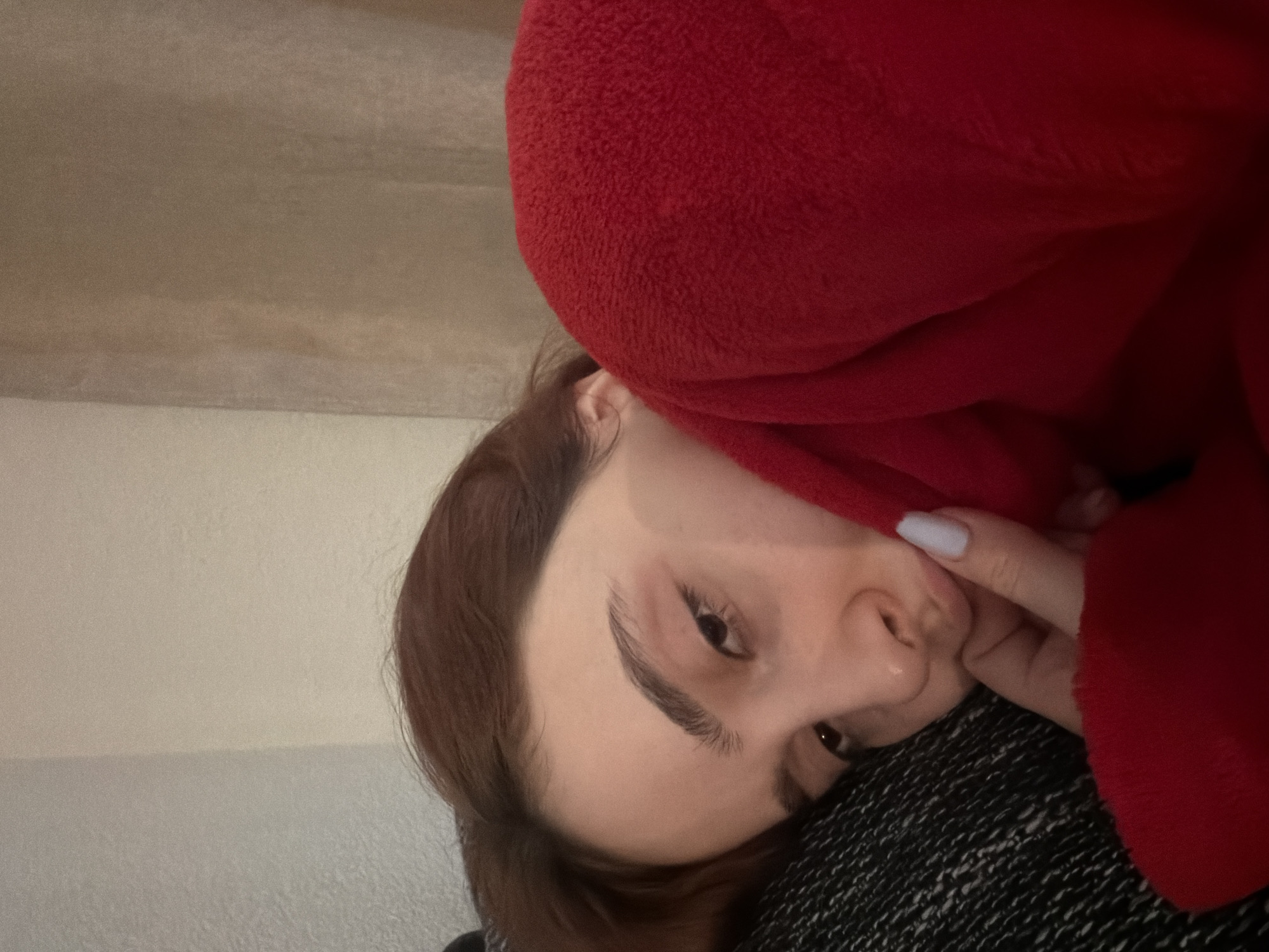 A woman is lying down, possibly in a residential setting. She is partially obscured by a red, plush fabric, possibly a blanket or hoodie, which covers part of her face. One hand with a white nail polish is visible, touching the fabric. Her brown hair is visible, and she is looking towards the camera. The background shows a beige-colored wall. The location in the city of Strasbourg, France, cannot be determined from the provided image.