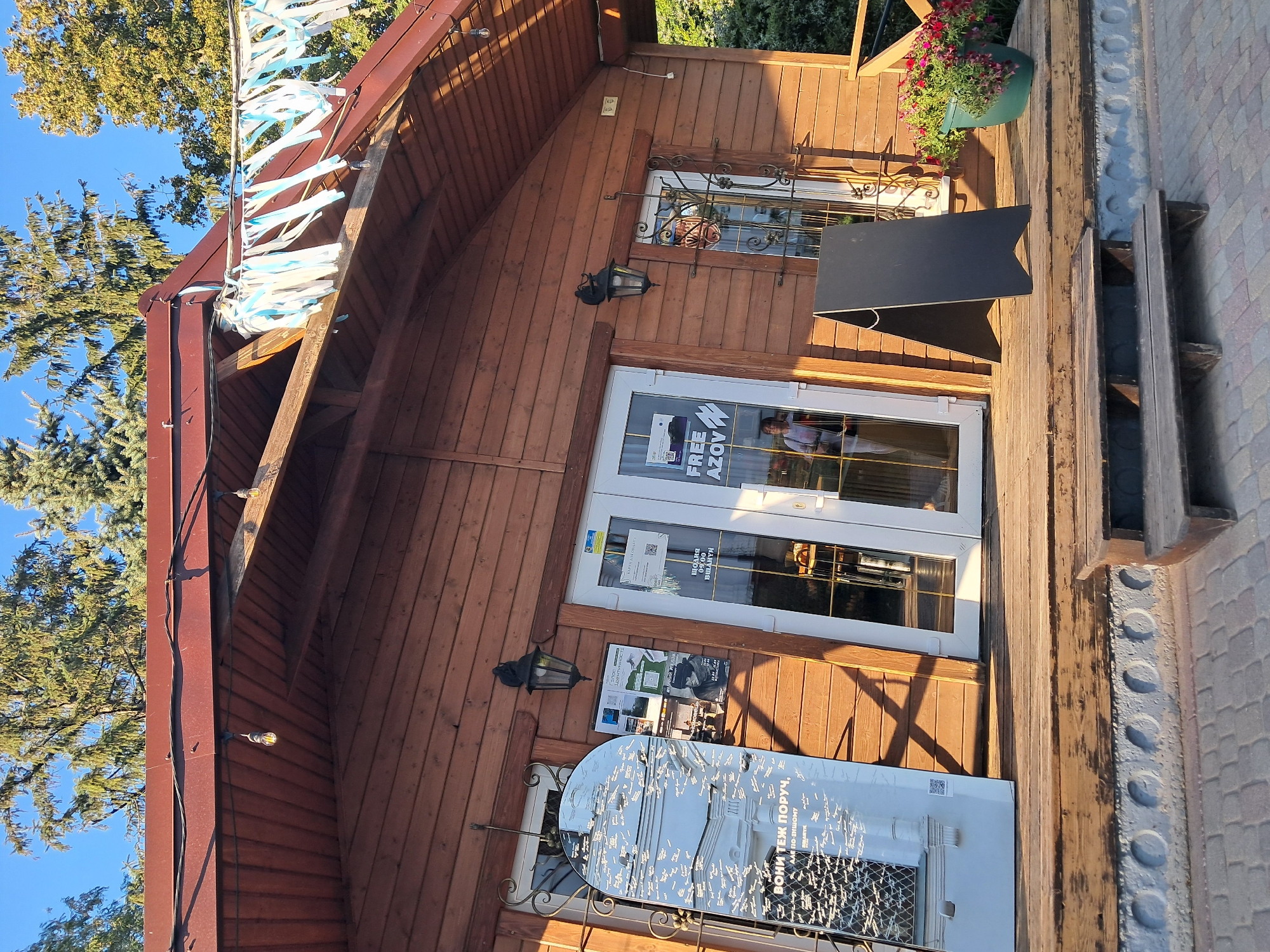 The image shows the exterior of a wooden building with a red-brown roof. The building appears to be a small shop or cafe. There are double glass doors in the center of the building, and a mirror is leaning against the left side. A black A-frame sign stands on the wooden porch in front of the building. In front of the sign, there is a green pot with red flowers. There is a wooden bench in front of the porch, and a brick walkway. The sky is blue, and there are trees in the background. The building is angled, suggesting the photo was taken from a low angle or the ground is uneven.