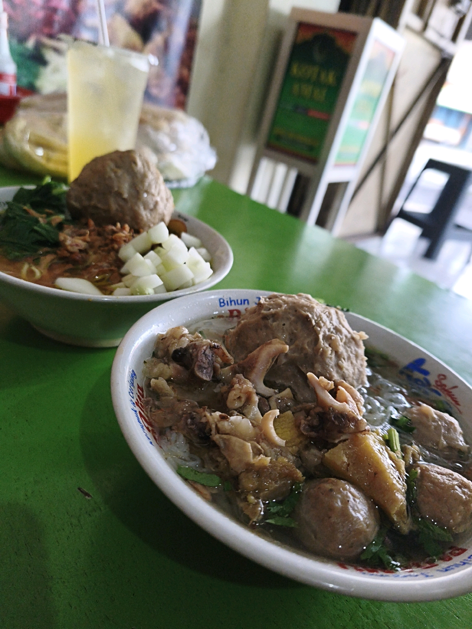 Two bowls of Indonesian meatball soup (bakso) are presented on a green table in what appears to be a casual dining establishment. The bowl in the foreground contains large meatballs, pieces of meat, and noodles in a clear broth, with some greenery. The bowl further back has noodles, greens, diced white vegetables, and a single large meatball. To the left, a glass of yellow liquid with ice and a straw is visible, along with a plastic bag. In the background, a blurry sign with text and graphics can be seen, suggesting a street-side eatery. The overall lighting suggests an indoor setting with ambient light. The restaurant appears to be located in the Selokaton area of Indonesia, evidenced by the visible text on the bowl in the foreground which reads "Bihun 7 Selokaton".