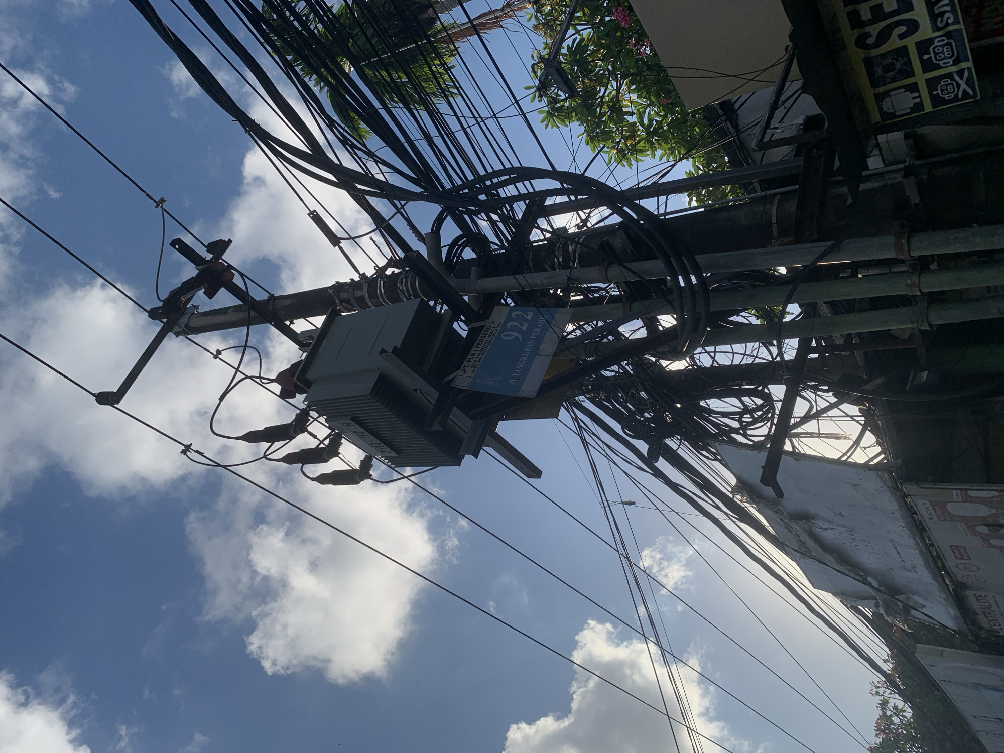 This media file is a low-angle shot looking up at a utility pole laden with a dense network of electrical wires, set against a bright blue sky with fluffy white clouds. The perspective is disorienting due to the pole's vertical orientation in the frame.
Dominating the scene is the complex web of black cables, some thick and others thin, bundled together and secured to the pole with various attachments. A grey electrical transformer box is visible, mounted on the pole. A blue sign is attached to the transformer box, displaying text in Indonesian. Part of the text reads "PT. PLN (PERSERO)" and "JL TANGKUBAN PERAHU", identifying it as belonging to the state-owned electricity company and indicating a street name. Another sign, yellow with black text and icons, is partially visible on the right side of the frame, with the word "SE" prominently displayed.
Lush green foliage with some pink flowers is visible at the top of the frame, suggesting the presence of trees or plants. The sunlight is strong, casting shadows and highlighting the textures of the wires and pole.
The overall impression is one of urban infrastructure, likely taken during the daytime, possibly in the morning or afternoon given the bright sky and sunlight. The dense arrangement of wires suggests a busy electrical distribution point. The visible text indicates a location in Indonesia, specifically near Tangkuban Perahu street, which is in the North Kuta area. No people are visible in the frame.