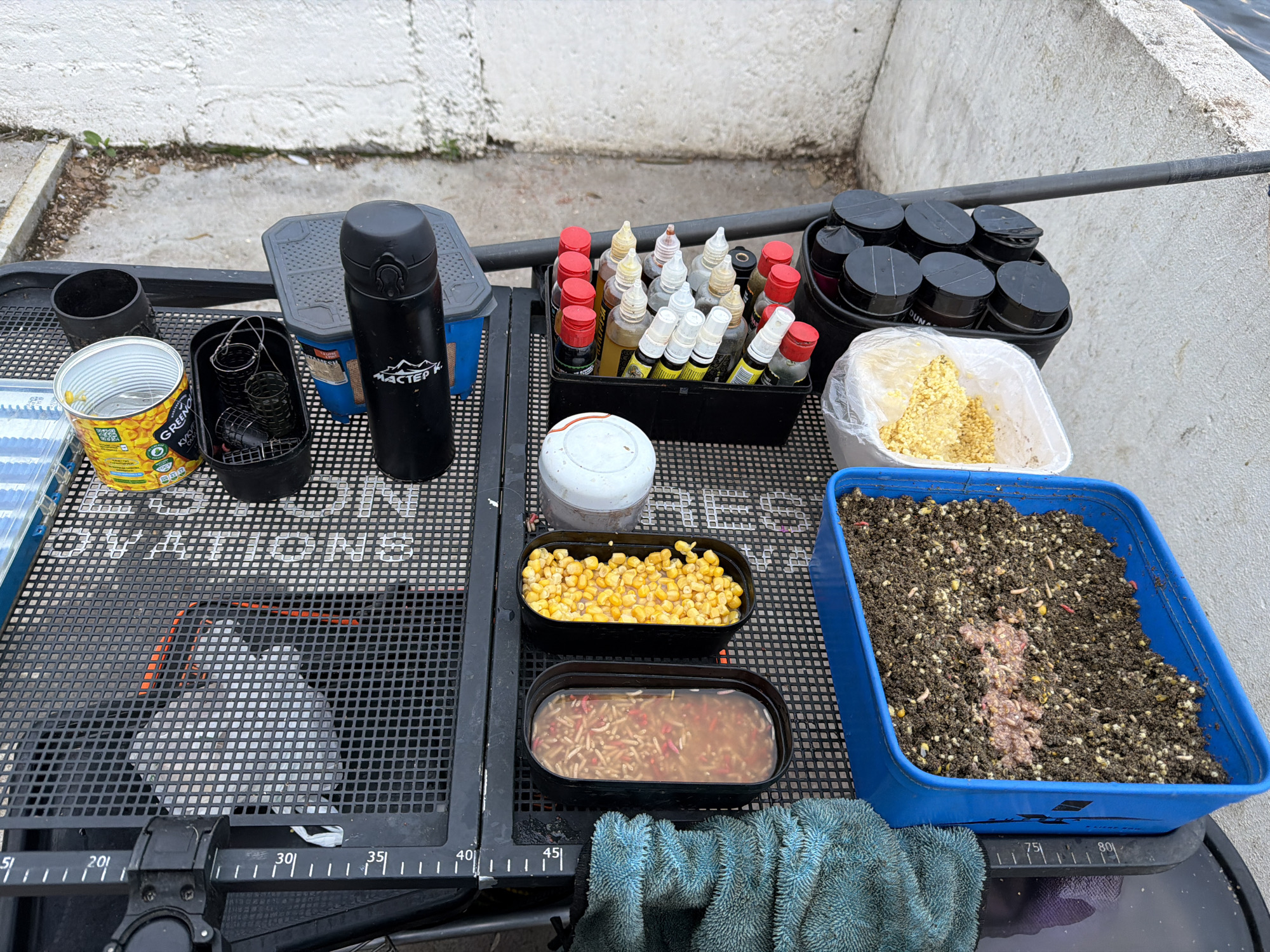 This image captures a fishing setup laid out on a black grid-patterned side tray next to a concrete wall and water in the city of Chaikovsky, Russia. The tray features various items, including an open can of Greenfoods sweet corn, a black container holding several metal fishing feeders, and a black insulated bottle with a "МАСТЕР К" logo. To the right, a black caddy contains multiple small bottles with red or white caps, likely fishing attractants or dips. Further right, another black container holds several round, black-lidded tubs. A white plastic container with a clear plastic bag holds yellow granular feed, while two black rectangular containers display prepared bait: one with sweet corn kernels in liquid, and another with a mixture of live maggots (some red) in liquid. A large blue rectangular container is filled with dark groundbait mixed with corn and maggots, with a central mound of pinkish, ground-up bait. A blue towel is draped below the blue bait container. The background shows a weathered concrete wall, and a glimpse of calm water is visible on the upper right side. Ruler markings are present along the edge of the fishing tray. No individuals are visible in the scene.