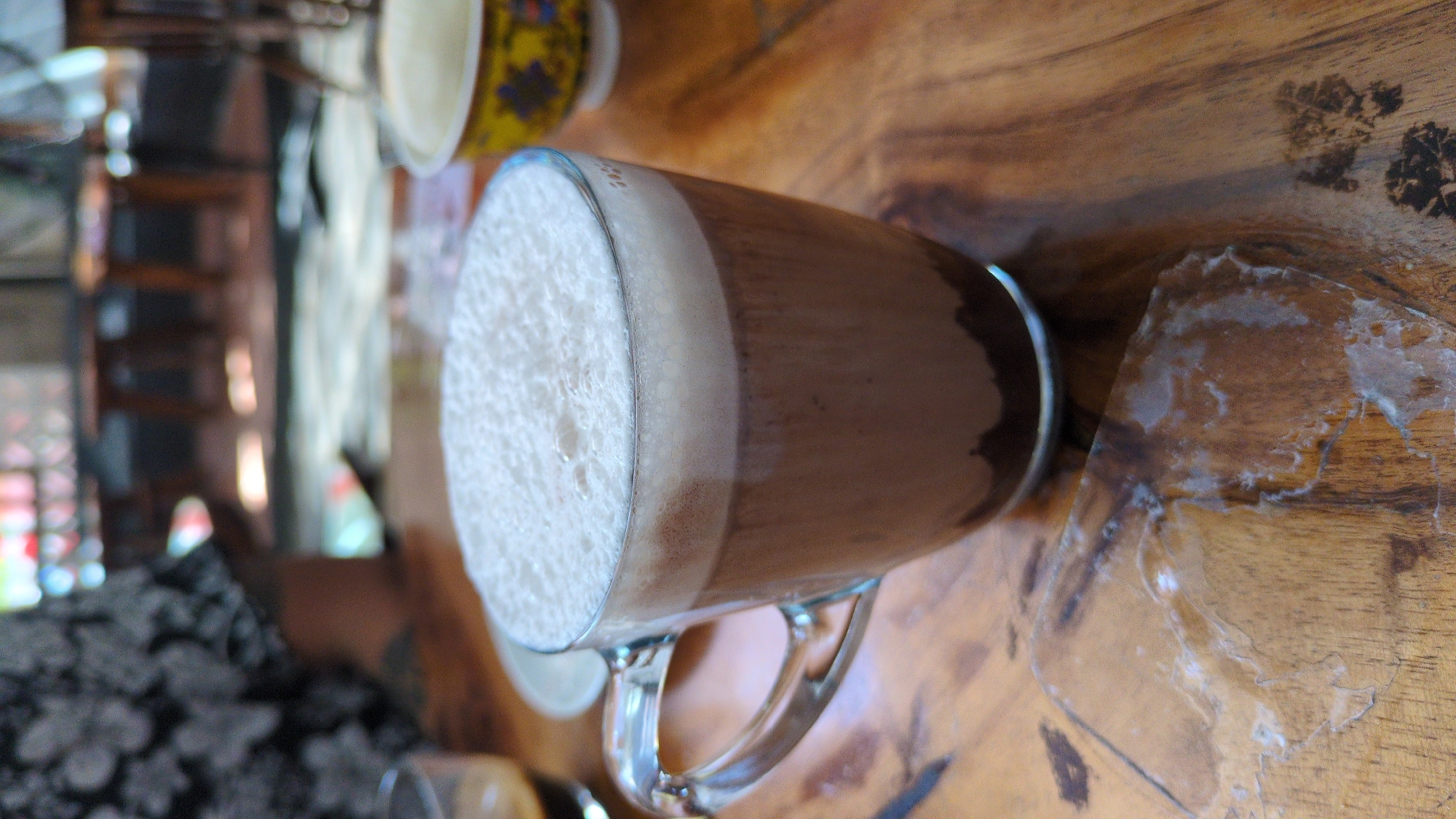 The scene is set indoors on a wooden table in Quy Nhon Ward, Vietnam. A clear glass mug, featuring a handle on the left, is centrally positioned. It contains a dark brown liquid, approximately two-thirds full, topped with a dense layer of white foam. To the upper left of the mug, a small clear glass holding a dark liquid is partially visible. On the wooden table surface, in the lower-left foreground, lies a piece of clear, crinkled plastic with white residue adhering to it. The wooden table exhibits visible grain patterns. In the blurred background, to the left, the torso and arm of an individual wearing a dark shirt with a light-colored floral or abstract pattern are visible. To the right in the background, a yellow bowl with a blue floral design is partially discernible. Further elements in the out-of-focus background include multiple dark wooden stools and light-colored flooring, indicative of a cafe or restaurant interior.