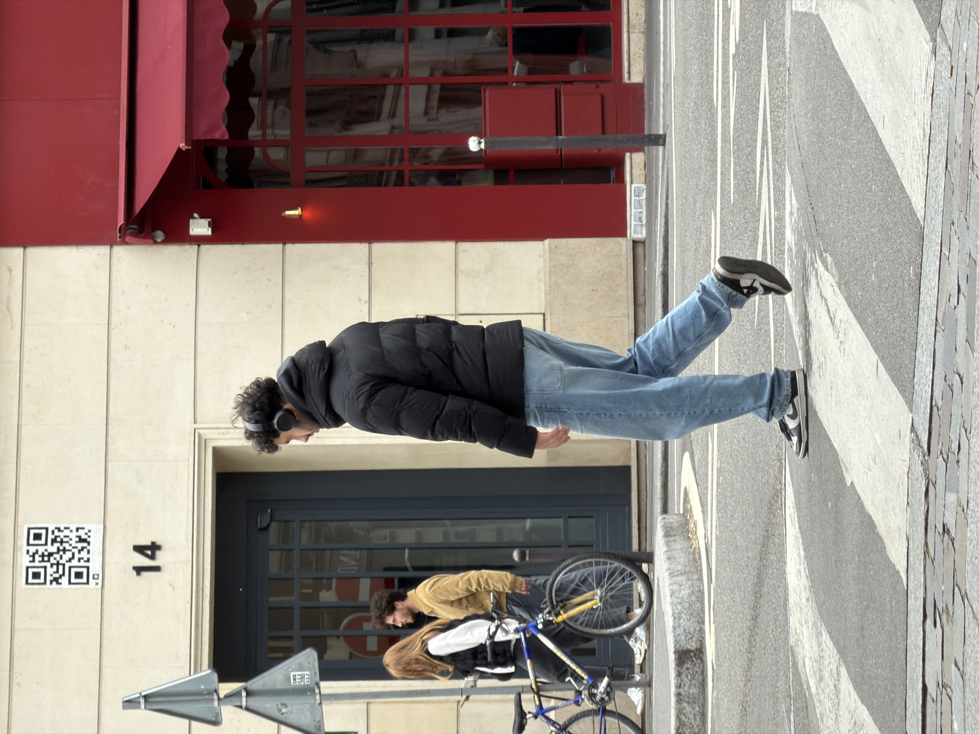 A street scene in Paris, France, shows a man in a black puffer jacket, blue jeans, and sneakers walking on a sidewalk with white stripes. He is wearing headphones. Behind him, another person stands beside a bicycle. The address on the building is marked "14." A QR code and street sign are on the building as well. The building has a red awning and window frames. The sidewalk is made of grey stones.