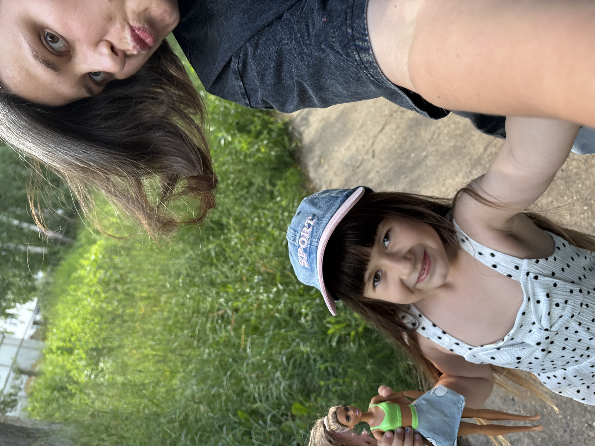 In this selfie, two females are featured outdoors, with a focus on their faces and upper bodies. The person in the foreground on the right is a young girl with dark hair, bangs, and brown eyes. She is wearing a blue denim cap with pink trim that has the word "SPORT" embroidered on it in white letters. She is also wearing a white dress with black polka dots. Her expression is a cheerful smile. In her hands, she holds a Barbie doll wearing a green top and a light blue skirt.
Partially visible in the upper left is the face of an adult female, also with dark hair. She is making a kissing face towards the camera. She is wearing a dark gray t-shirt. The background consists of lush green grass and a paved walkway. The lighting suggests it is daytime, likely sunny given the bright colors. The setting appears to be a park or a residential area, possibly in Chaikovsky, Russia, based on the provided location context. The overall mood is lighthearted and joyful, capturing a moment between an adult and a child enjoying an outdoor activity.