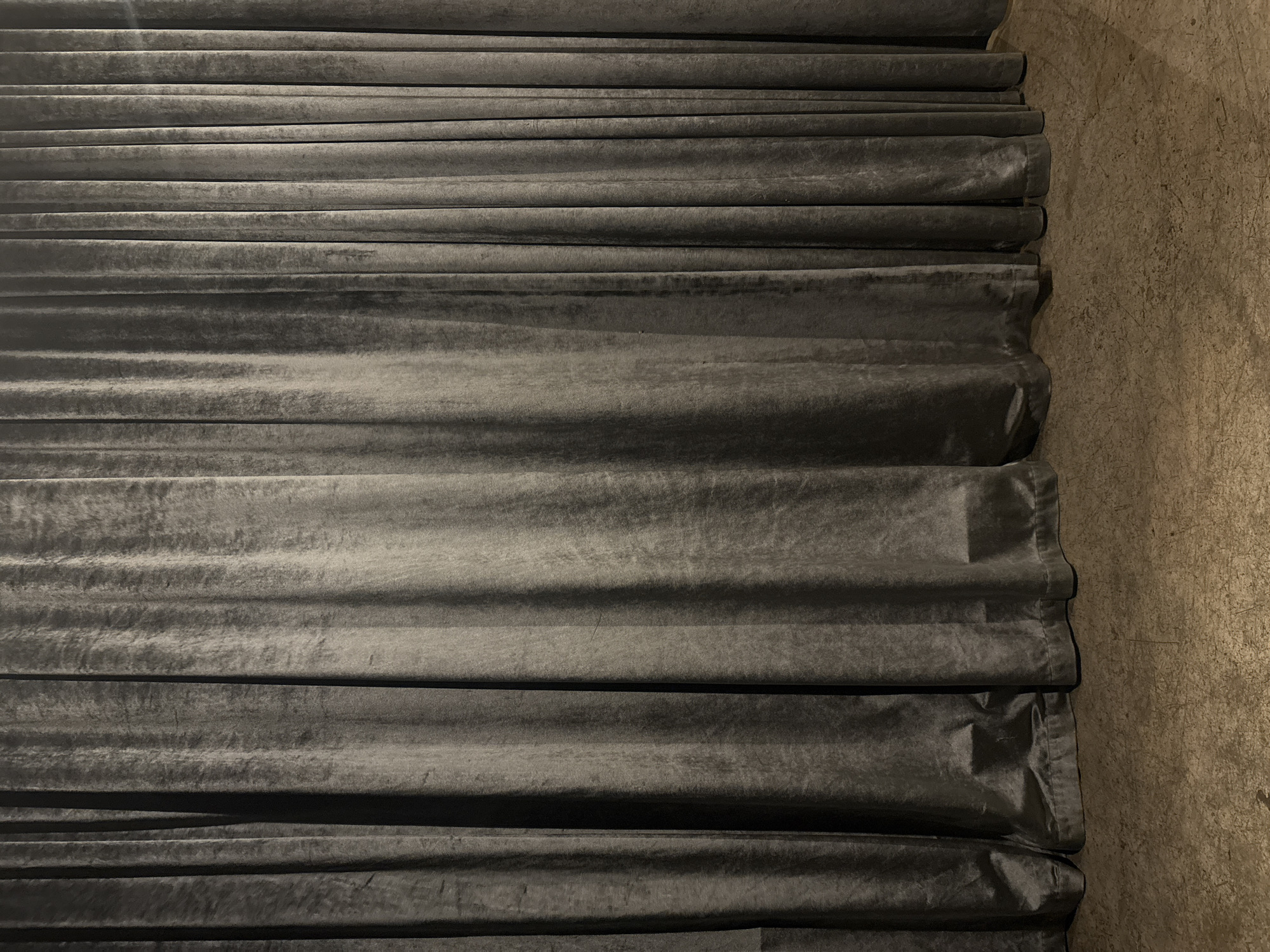 The image depicts a close-up, eye-level view of a dark gray, velvet curtain. The curtain is tightly gathered, creating numerous vertical folds and pleats that run from top to bottom. The texture of the velvet is evident, with subtle variations in light and shadow highlighting the fabric's plushness. At the bottom of the curtain, the hem is visible, resting on a textured floor. The floor appears to be made of concrete or a similar material, with a mottled brown and gray appearance and visible scuff marks or imperfections. The lighting is dim, casting shadows that deepen the folds of the curtain and illuminate the floor with a warm, indirect light. There are no people or discernible objects other than the curtain and floor. The scene is consistent with a theater, stage, or an event space where curtains are used for ambiance or to conceal a backdrop. Given the prompt's specificity, this scene is located in the city of Burgas, Bulgaria.