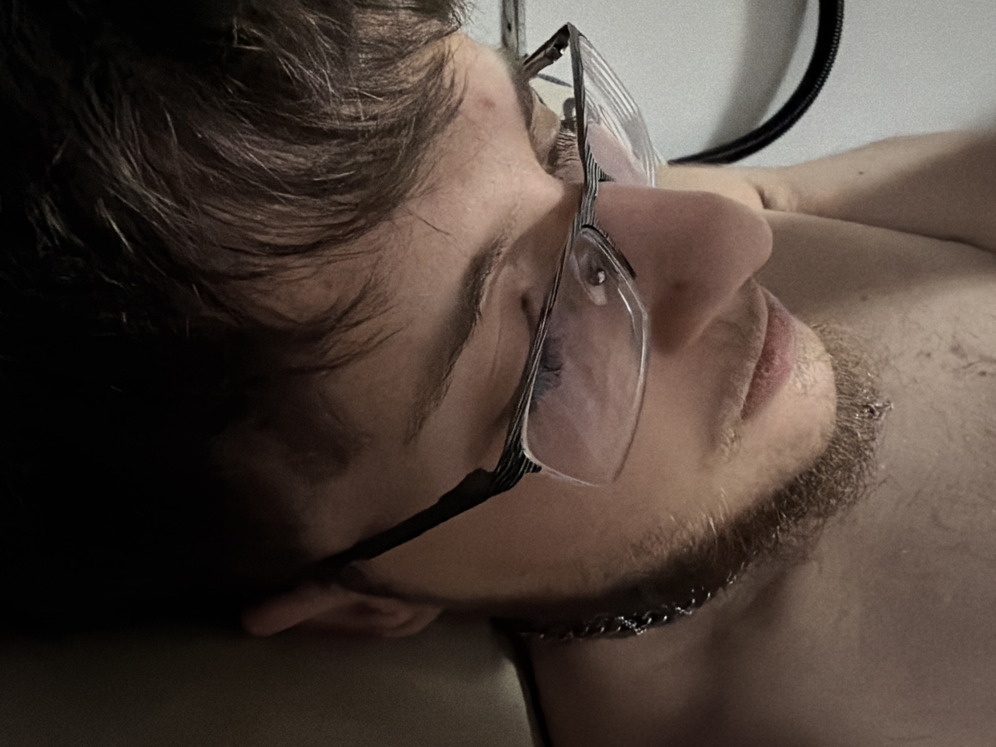 The image shows a close-up, low-angle shot of a person lying on their side, with their head turned to the side and partially obscured by their hair. They are wearing eyeglasses with thin metal frames and are looking down. The person has short, dark hair and a light beard and mustache. A thin, silver-colored chain necklace is visible around their neck. The background is a plain, light-colored wall with a black corrugated cable attached near the top right. The person's skin tone appears fair. The lighting is dim and casts shadows, creating a moody atmosphere. No specific location in Plovdiv, Bulgaria, can be determined from the visual information provided.