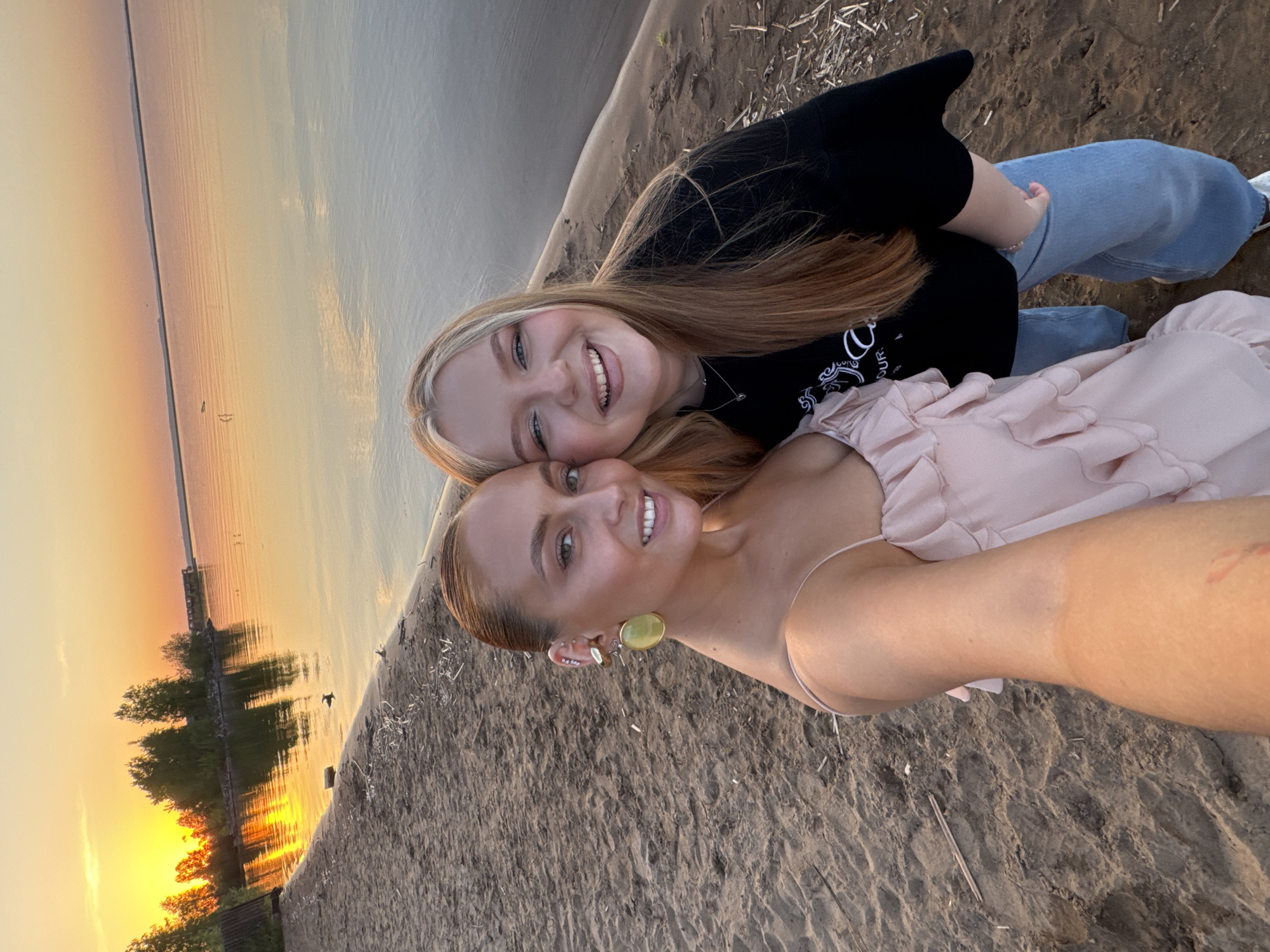 Two young women are lying on a sandy beach at sunset in Chaikovsky, Russia. One woman has blonde hair, is wearing a black shirt and blue jeans, and is positioned behind the other woman, who has auburn hair and is wearing a light pink dress. Both are smiling at the camera. The background features the Kama River at sunset. Trees and a pier are silhouetted against the setting sun.