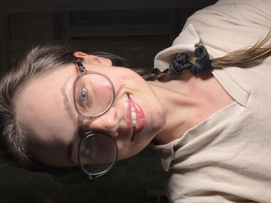 The image depicts a young adult female in an indoor setting within Scafati, Italy. She is positioned from the chest up, centered in the frame, and looking directly at the camera with a subtle smile, revealing her upper teeth and glossy pink lips. Both cheeks display dimples. She has light skin and brown hair, styled in a single braid draped over her right shoulder (viewer's left), secured with a black scrunchie. She wears clear-framed, semi-transparent eyeglasses; her left eye (viewer's right) is blue and clearly visible, while her right eye (viewer's left) is partially obscured by shadow and a reflection on the lens. Her attire consists of a light beige or cream-colored knit polo-style shirt featuring a ribbed collar and a V-neck placket. The environment is dimly lit, with a distinct light source illuminating the right side of her face (viewer's left), creating a strong shadow across the left side of her face (viewer's right). The background is dark and indistinct, with a faint rectangular outline, possibly a window or door frame, visible on the far right.