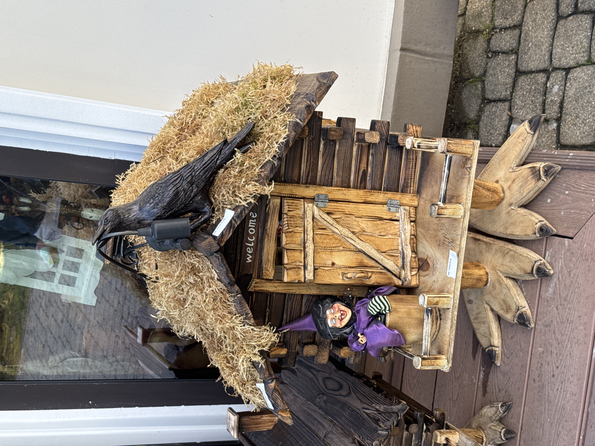 A close-up, slightly high-angle shot shows a Halloween decoration featuring a wooden birdhouse-like structure with a black crow perched on the roof. The roof is covered in dried moss-like material. A small wooden door is visible with a welcome sign. Below the door, a miniature witch figurine with a purple outfit and a pointed hat is positioned. The base of the structure has wooden accents resembling claws or talons. The backdrop includes a window and a stone-paved area, suggesting an outdoor setting. There is no indication of the image being located in Sirius, Russia.