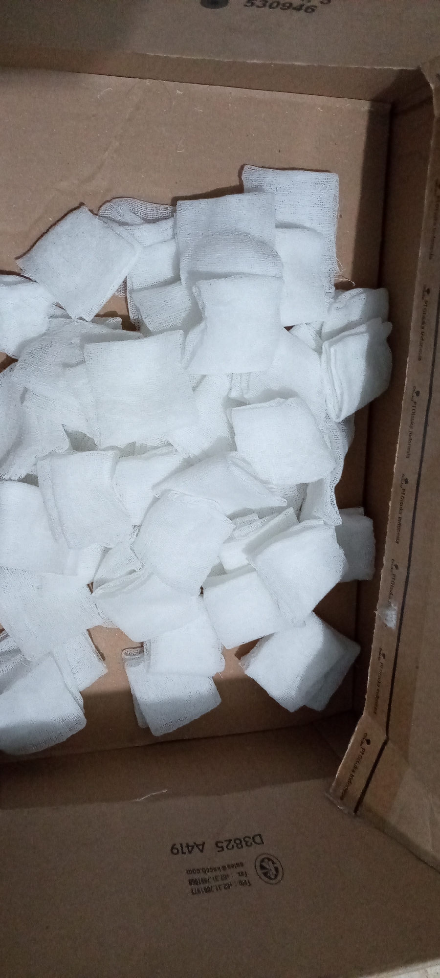 The image displays a close-up, top-down view inside an open cardboard box filled with numerous white, square-shaped gauze pads. The pads are loosely piled, occupying most of the box's interior space. The box itself is made of light brown cardboard, with visible printing on its inner walls.
There is no activity taking place; the image is a static shot of the contents of the box. The lighting appears to be artificial, common for an indoor setting, with no indication of natural light or specific time of day.
Notable details include the material of the pads, which appear to be medical-grade gauze, suggesting the box contains supplies for healthcare or first aid. The cardboard box bears several printed texts, some of which are partially obscured or repeated. Visible text includes "530946", "PT Otsuka Indonesia" (with slight variations like "PrOtsuka udonesia", "Prorsuka ndonesia", "ProDuka indonesia", "Pt Otsuka Indone"), "sales@ksccb.com", "Fax: +62.31.7851968", "Telp: +62.31.788197", and "D3825 A419". These details, particularly the company name "Otsuka Indonesia" and the Indonesian telephone/fax codes (+62), strongly suggest the box originated from or is associated with Indonesia, aligning with the provided location context of Lhokseumawe, Indonesia.
