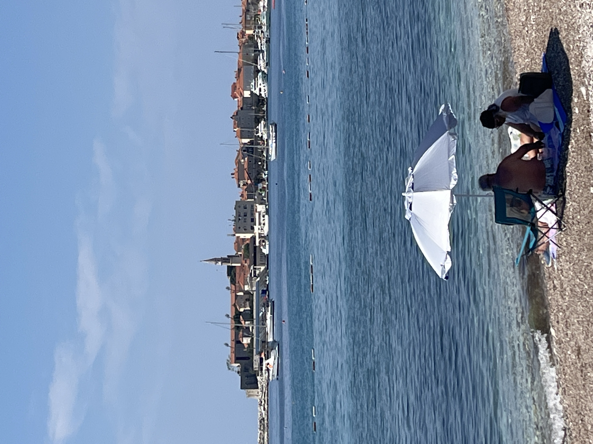 The image captures a coastal scene in Budva, Montenegro. A beach stretches along the right edge, with waves gently lapping the shore. Two individuals are seated on the beach, partially obscured by a white umbrella. In the distance, a row of buildings lines the coastline, with a mix of residential and commercial structures visible. The water between the beach and the buildings is calm and blue, with a few boats anchored in the distance. The sky above is clear and pale blue, with a few wispy clouds.