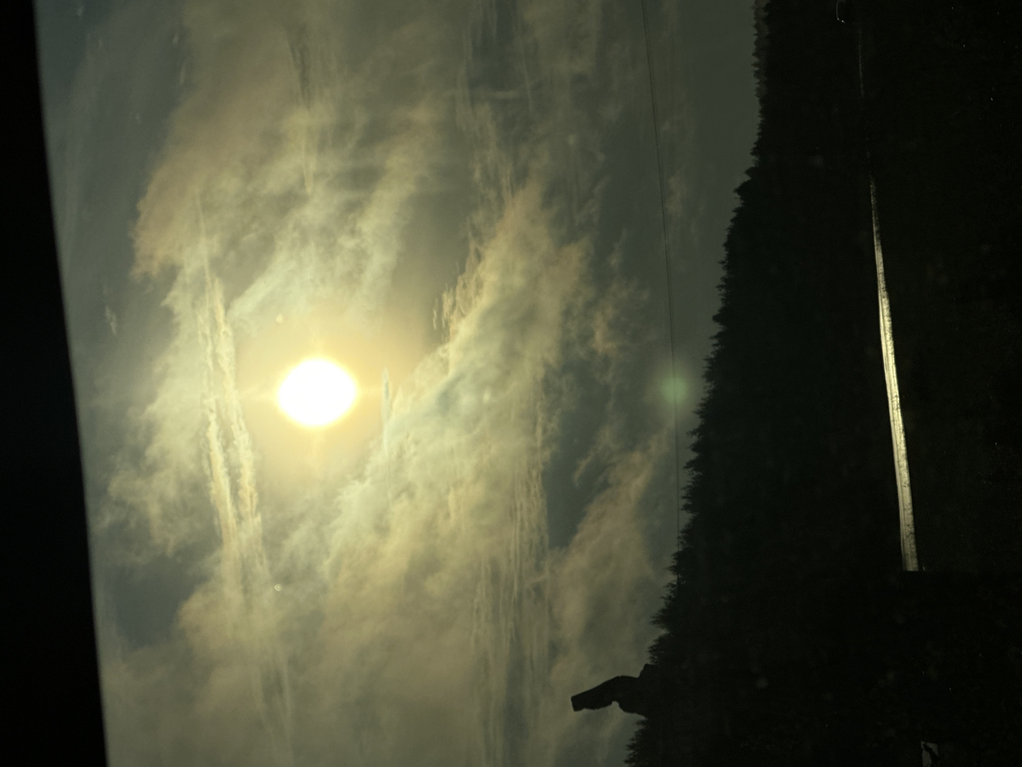 The image depicts a silhouetted figure standing on a dark, tree-lined hill against a dramatic, cloudy sky illuminated by a bright sun. The figure appears to be a statue or monument, with a distinctive angular shape. Below the hill, a dark road or surface reflects the light, suggesting it might be wet or metallic. The overall scene is dark and atmospheric, with the sun's glare dominating the upper portion of the frame. The specific location in the city of Яган-Докья, Russia, is not discernible from the image itself.