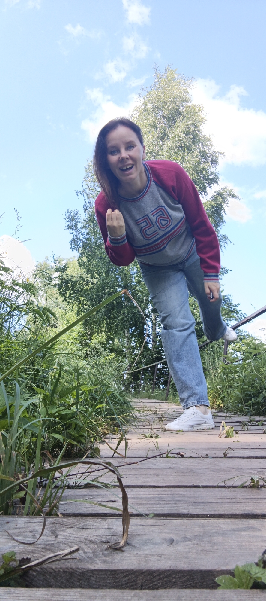 A young woman is captured from a low angle on a wooden boardwalk surrounded by lush greenery and a bright blue sky with scattered clouds. She is wearing a gray and pink sweatshirt with red and blue trim and blue jeans, and white sneakers. The woman is leaning forward, looking towards the camera with a wide smile and a raised fist, as if in celebration or excitement. She appears to be walking on the boardwalk. The scene is set outdoors, possibly in a park or natural area, and the wooden boardwalk suggests it could be a nature trail or recreational path. The presence of trees indicates a natural, possibly rural or suburban environment. The lighting suggests it is daytime, with clear weather. The image is taken in Chaikovsky, Russia, where such boardwalks are common in natural parks and along the Kama River.