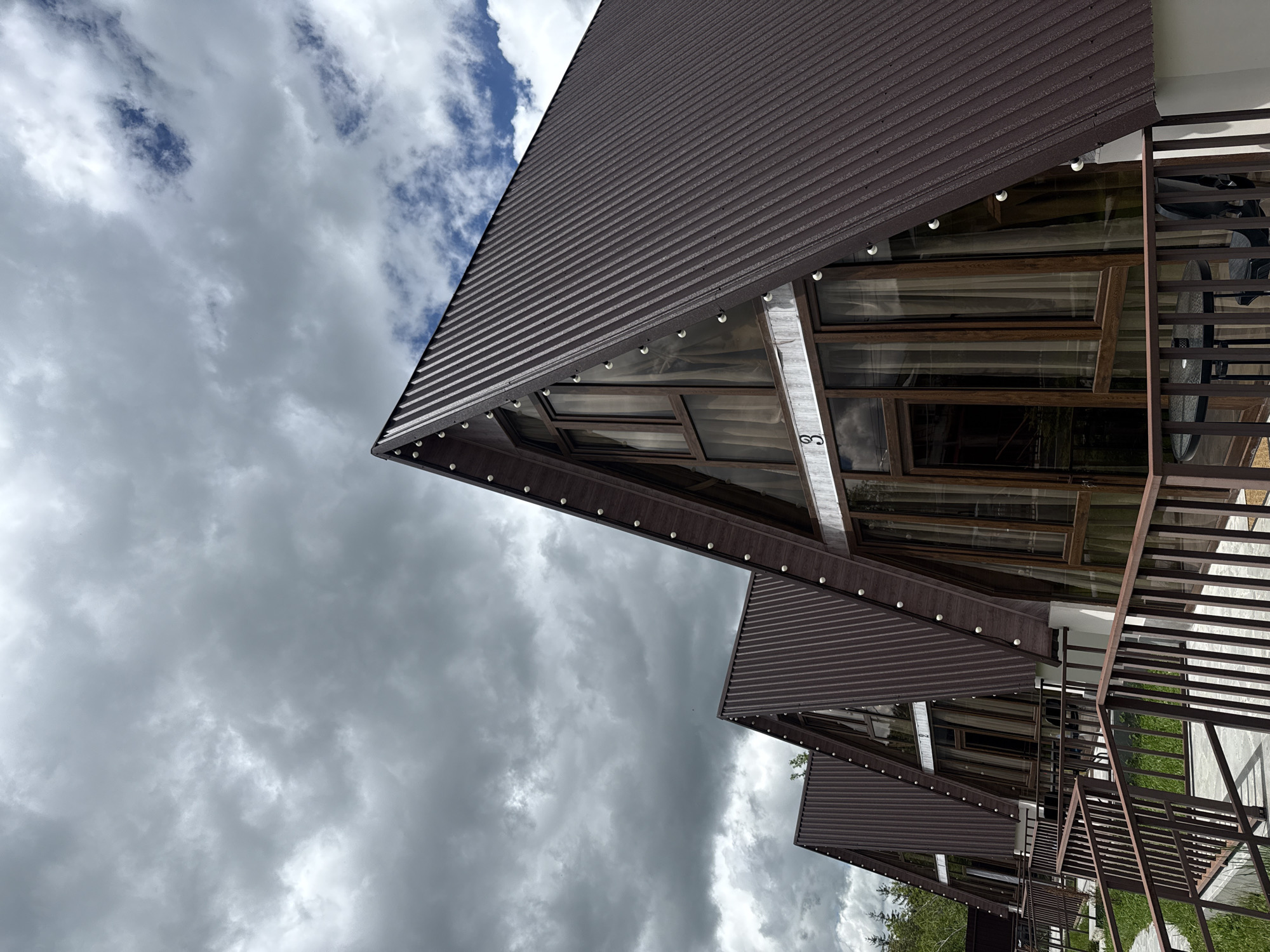 The image depicts the exteriors of two adjacent A-frame buildings, likely vacation homes or lodging, situated in Stepanavan, Armenia. The buildings feature brown, corrugated metal roofs and large windows framed with dark wood, allowing natural light to penetrate the interior spaces. A metal railing, matching the dark wood accents, runs along the exterior, indicating a balcony or elevated walkway. The sky above is overcast with dark, ominous clouds, suggesting an approaching storm or recent rainfall. The buildings appear to be nestled in a green landscape, with glimpses of trees visible through the railing.