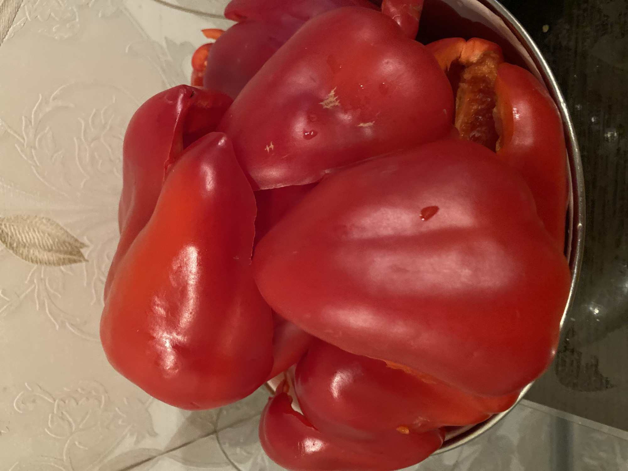 A close-up, eye-level, slightly downward-angled shot captures a metallic bowl overflowing with vibrant red bell peppers. The peppers are plump and glossy, with some showing slight indentations and water droplets on their skins. The bowl itself has a silver rim and is resting on a dark, reflective surface, likely a countertop. In the background, a light-colored wall with a subtle, embossed floral pattern is visible. The overall lighting is warm, highlighting the rich red of the peppers. There are no people present in the image. This scene does not provide any information that would allow for determining its location within Ivanteyevka, Russia.