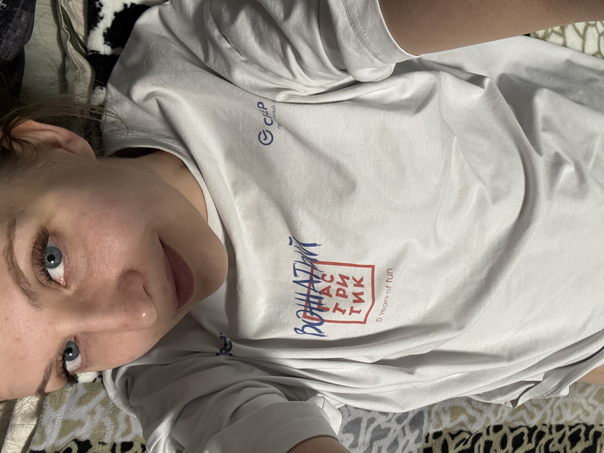 A woman is lying on a patterned surface, possibly a bed, wearing a white t-shirt with Russian text and logos on it. The text includes the phrase "Вощатный ТРИ ТИК" with "5 years of fun" written below. The logo on the upper left of the t-shirt is "CIRP technologies". The woman has light skin, blue eyes, and light brown hair. The location is likely Izhevsk, Russia, based on the Cyrillic text on the t-shirt.