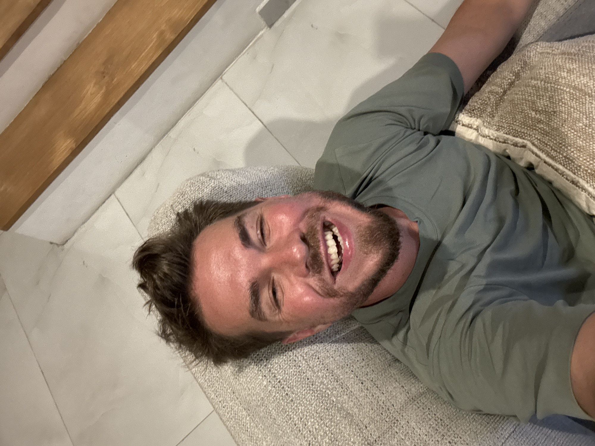 A smiling man with brown hair and a beard is lying down on a light grey cushion, possibly a part of a sofa or bed. He is wearing a light green t-shirt. The background consists of white tiled flooring and a light wooden structure. The image was most likely taken indoors. The location in North Kuta, Indonesia, is not verifiable from this image.