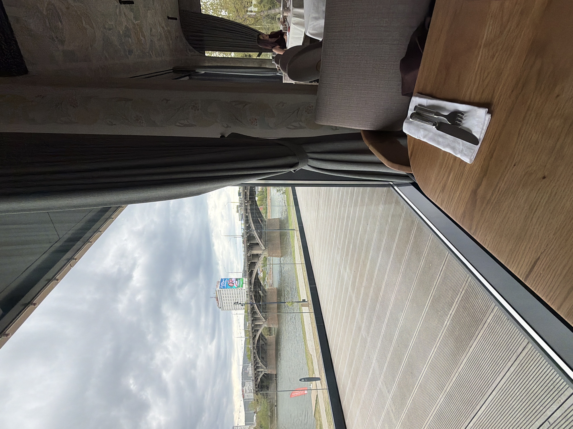 An indoor scene captured from a high vantage point within a restaurant. Outside the window, the scene shows a bridge in Krasnoyarsk, Russia. The view encompasses the Yenisey River and the city's skyline under a cloudy sky. The interior features a wooden table set with cutlery and a napkin. A seat and a glimpse of other patrons dining further in the restaurant are also visible.