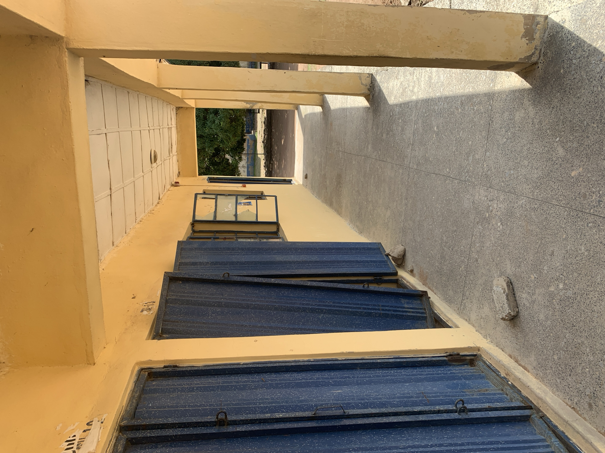 The image shows a sunlit outdoor hallway or veranda with a yellow-painted building on the left. The hallway has a concrete floor and is supported by beige-colored pillars on the right. On the left side of the building, there are two large blue metal doors, one of which is partially open. Above the doors, there are some small, faded markings on the yellow wall, including what appears to be "31-13". To the right of the doors, there is a window with multiple panes that is partially open. The hallway extends into the background, where trees and some scattered debris are visible. The scene appears to be located in an outdoor public space, possibly in a school or administrative building in Jalingo, Nigeria, given the architectural style and vegetation. There are no people visible in the image.