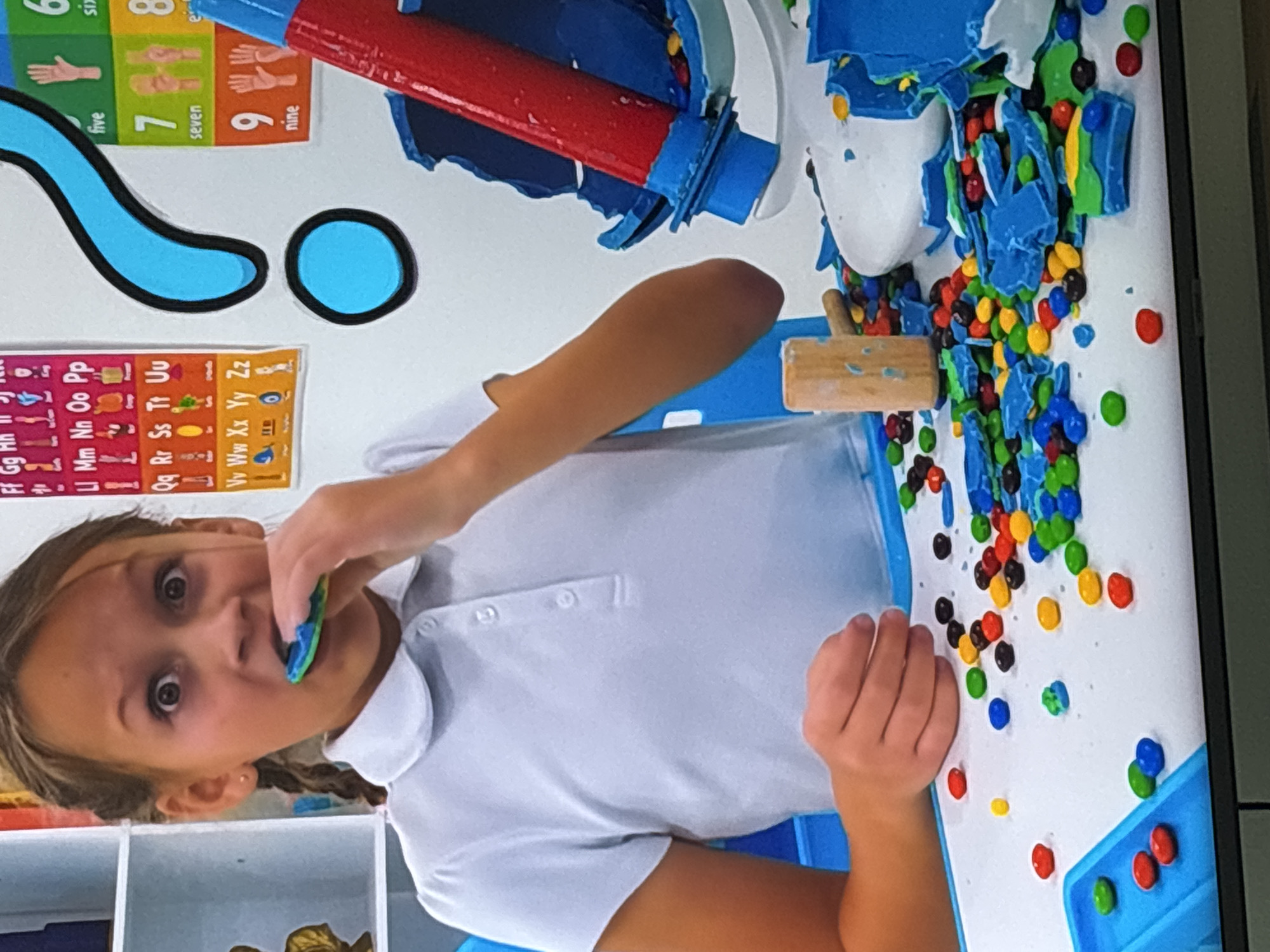 The image shows a young girl with wide eyes and an open mouth, holding a blue and green candy in her mouth. She is wearing a white polo shirt. She is sitting at a table covered in scattered colorful candies, broken pieces of blue and white material, and a small wooden hammer. Behind her, on a white wall, are educational charts with letters and numbers. A large blue question mark decoration is also visible on the wall. The scene appears to be an indoor setting, possibly a classroom or a play area, with blue plastic chairs and shelving units visible. There is no indication of the city of Dubai or the United Arab Emirates in the image.