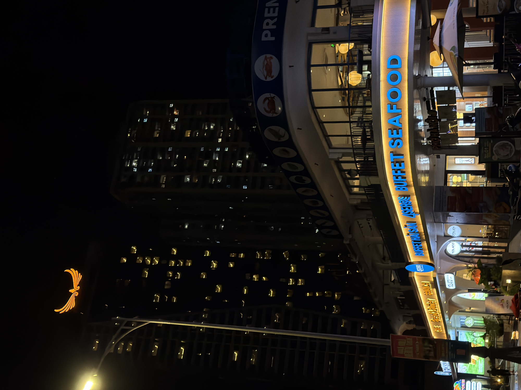 The image depicts a night scene in Đà Nẵng, Vietnam, featuring a multi-story building with illuminated windows. At the top of the building, a prominent golden phoenix logo is visible against the dark sky. The lower portion of the building houses a restaurant with bright blue and yellow neon signs. One sign reads "PREMIUM BUFFET THAI SEAFOOD," while another, in yellow, says "THE GREAT BUFFET BRUNCH." Below these, there are arched entrances to various establishments, including one with signage for "YOLATO" and "YOLATOY." Several people are present on the sidewalk in front of the restaurants. The street appears wet, suggesting recent rain. A street lamp illuminates the left side of the image. The architecture suggests a modern urban environment.