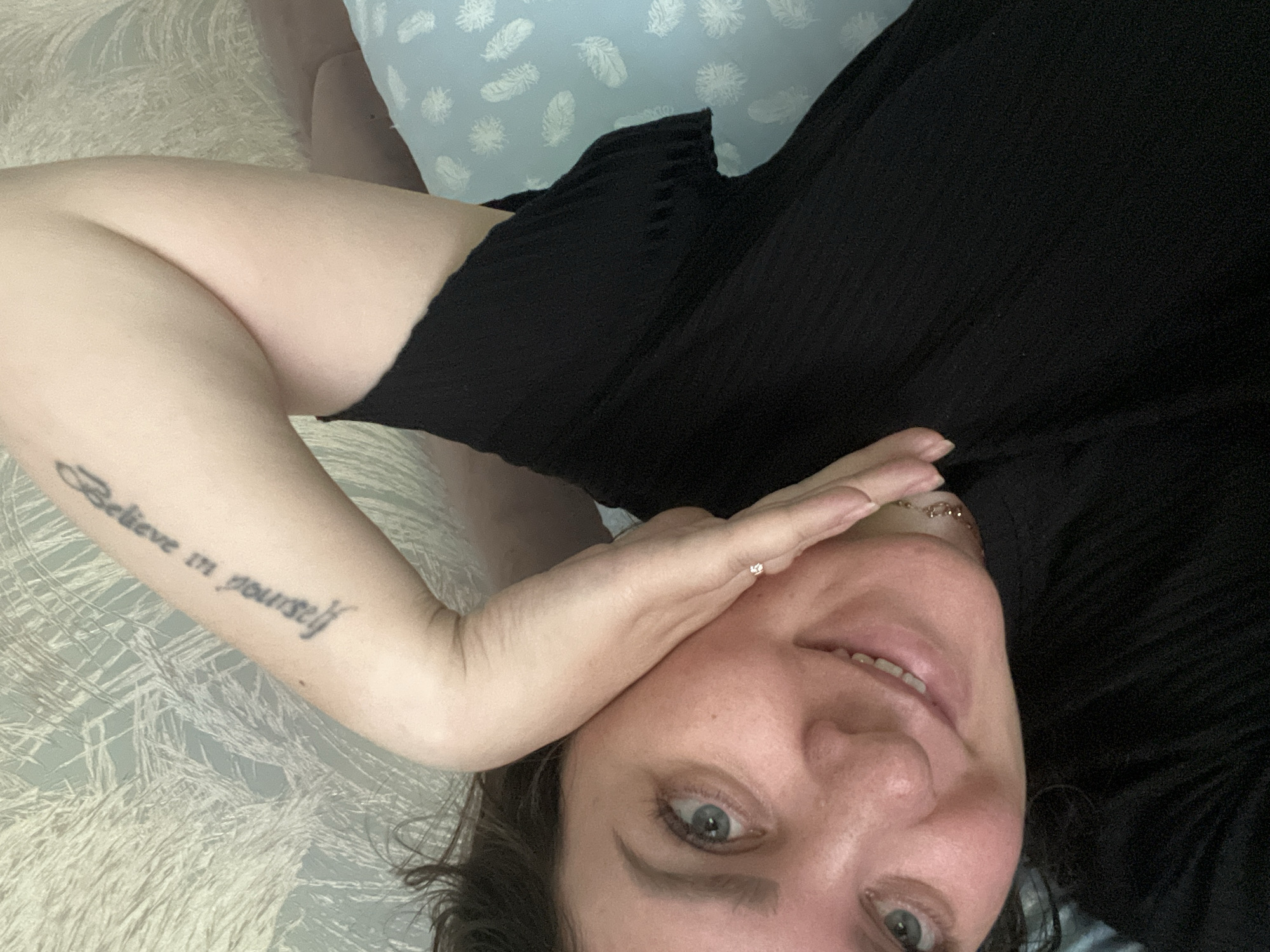 The image shows a close-up, high-angle, selfie-style shot of a woman lying down. She has light skin and blue eyes, with dark hair. She is wearing a black top and a delicate gold necklace. Her left arm is bent, with her hand resting on her cheek, and a tattoo reading "Believe in yourself" is visible on her upper arm. Her right arm is not visible. The background is a textured wallpaper with a light, muted pattern, and a light blue pillow with white feather designs is visible on the right. The scene appears to be an indoor setting, likely a bedroom, and no specific location in Noviy, Russia, can be determined from the image.