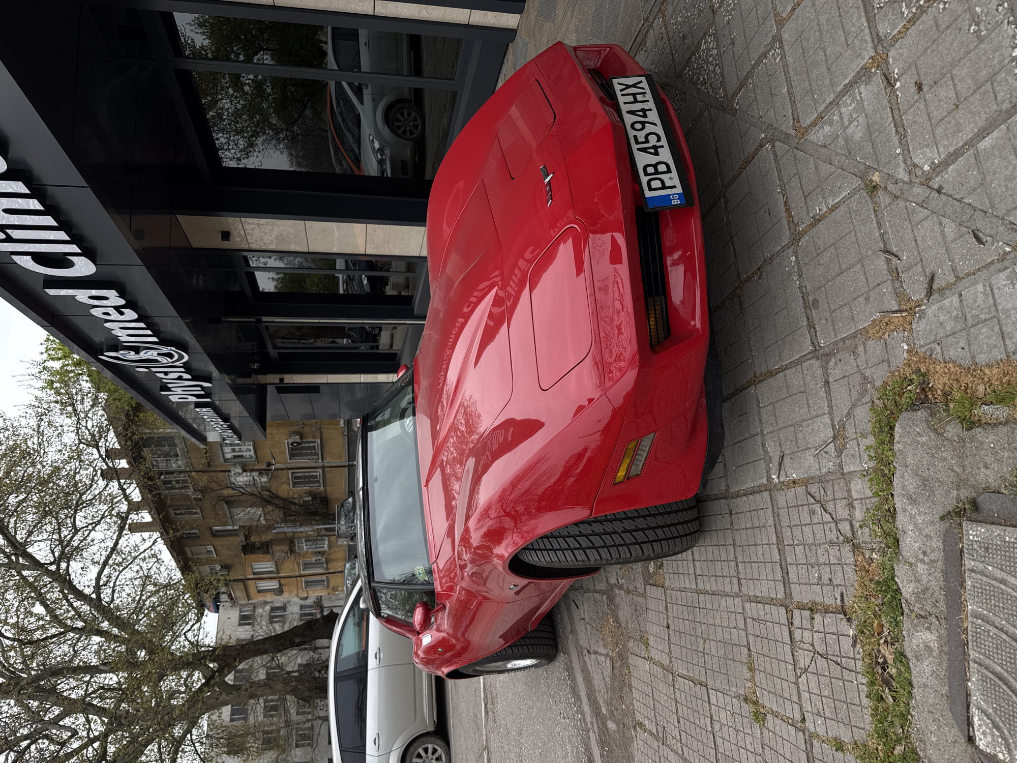 A red Corvette convertible is parked in front of the "Physio S Med Clinic" in Plovdiv, Bulgaria. The license plate reads "PB 4594 HX" and has the Bulgarian flag displayed. The car sits on a sidewalk paved with grey rectangular tiles, some with weeds growing in the cracks. Next to the Corvette is a silver minivan. A bare tree is visible on the left, in front of a multi-story building with apartments. The clinic's signage is large, black, and white. Reflections of other vehicles and foliage are seen in the clinic's glass facade.
