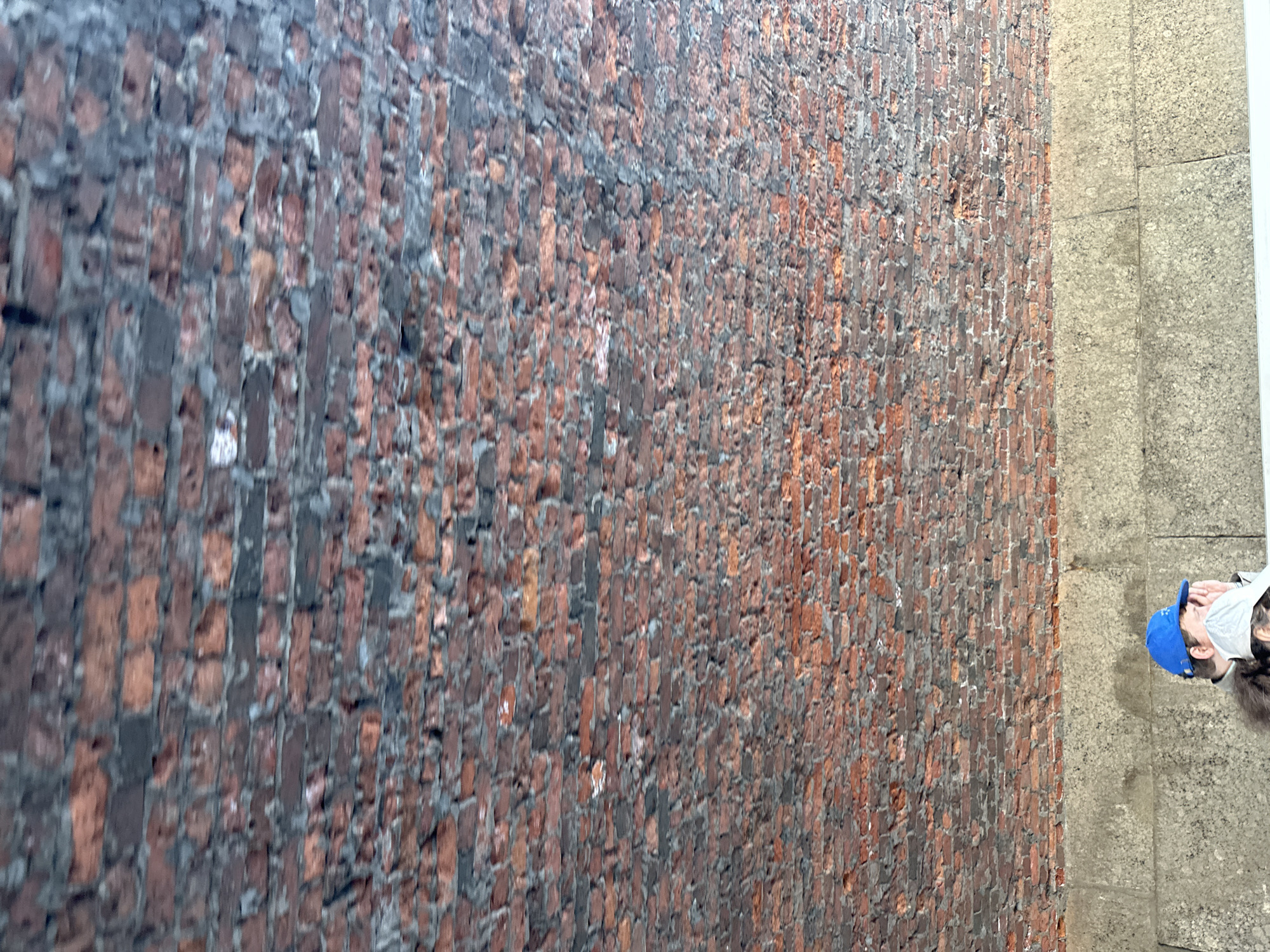 The image shows a brick wall next to a concrete walkway. A person wearing a blue baseball cap and a white face mask is standing on the walkway, with their back partially facing the camera. The wall is made of dark red bricks with some black staining, possibly from soot or moisture. The scene seems to be located in an urban environment.