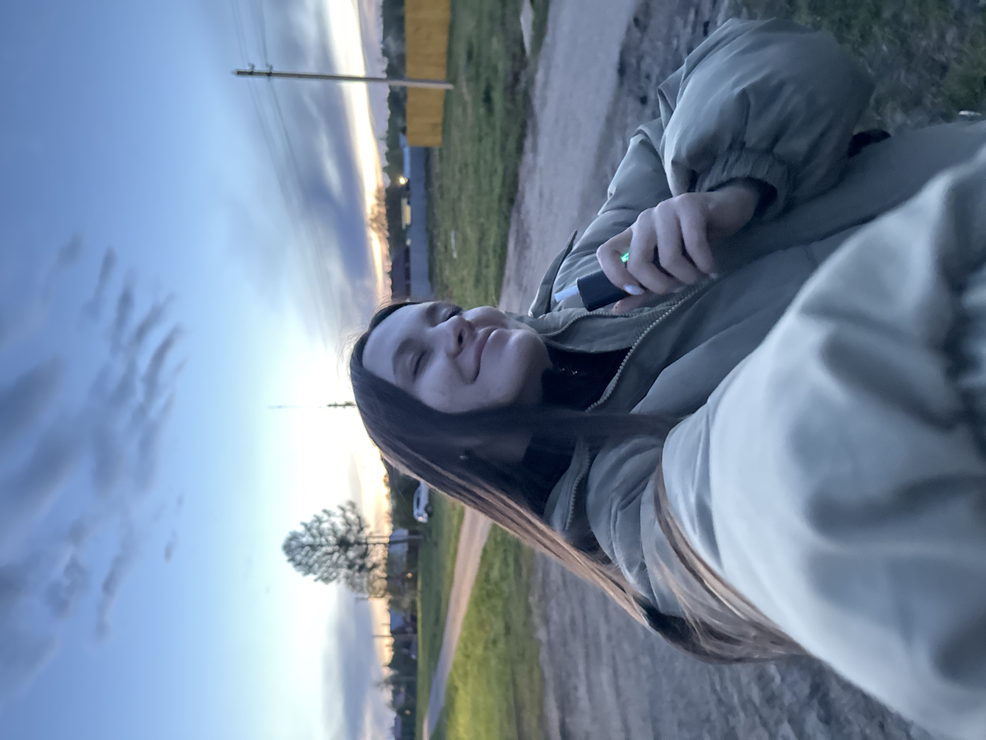 A young woman is holding an object that appears to be a vape pen, which is lit. She is wearing a puffy jacket and standing on a dirt road. The setting appears to be rural or semi-rural, as there are fields and trees in the background. In the distance, there are some buildings and utility poles. The sky is light, indicating it is dusk or dawn. It is not possible to determine the location of Красный Маяк, Russia from the image.