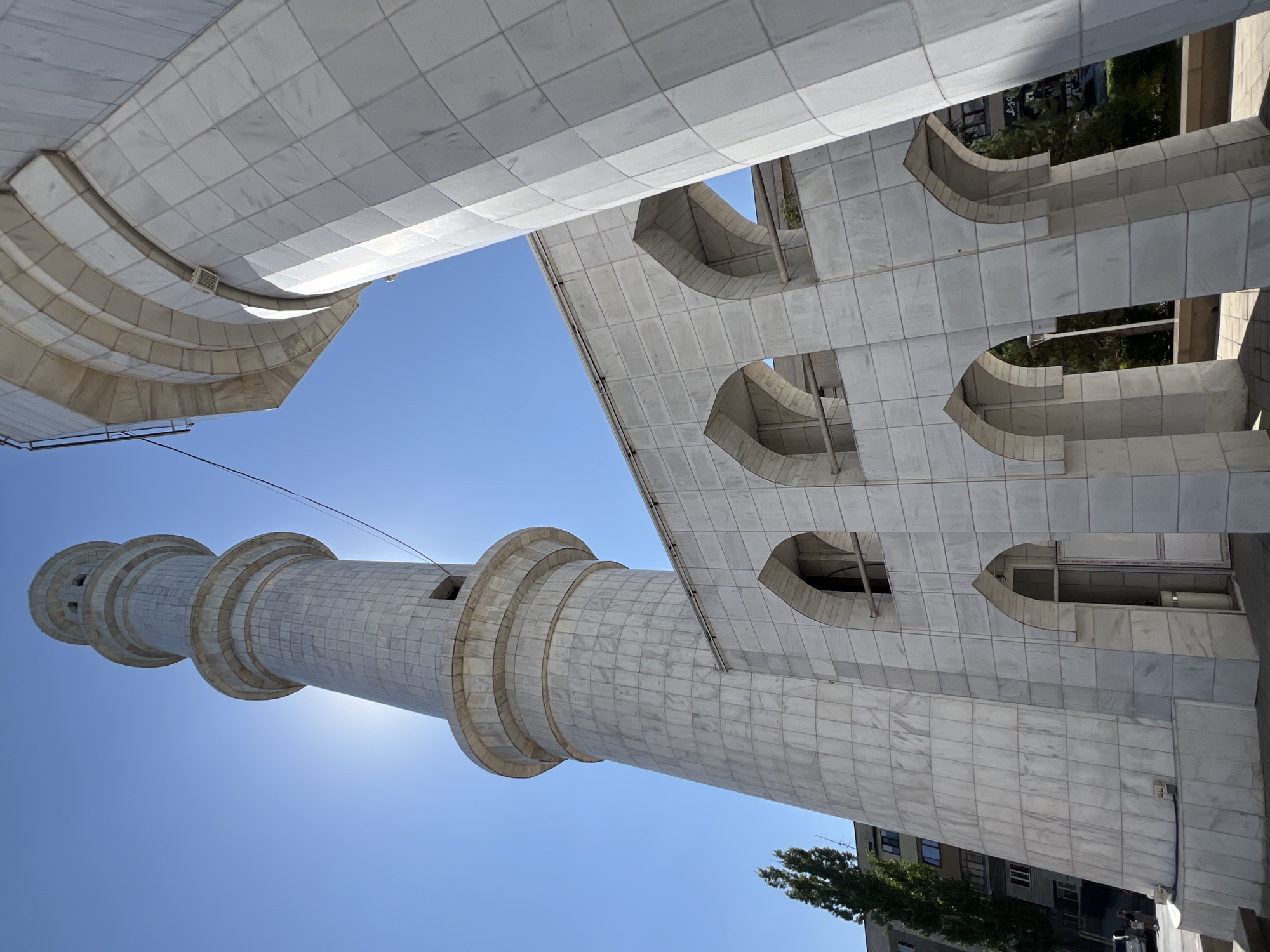 The image shows a wide-angle view of a modern mosque minaret and part of its adjacent building in Almaty, Kazakhstan, under a clear blue sky. The structure is primarily made of white marble tiles. The minaret is tall and cylindrical, tapering upwards with several horizontal bands. The main building has a facade with arched windows and openings. Sunlight is a strong element, creating lens flare behind the minaret. In the background, a tall, leafy tree and a multi-story building are visible, suggesting an urban environment. The perspective is from a low angle, emphasizing the height of the minaret and the architectural details of the mosque. The scene is static, with no visible human activity or interaction.