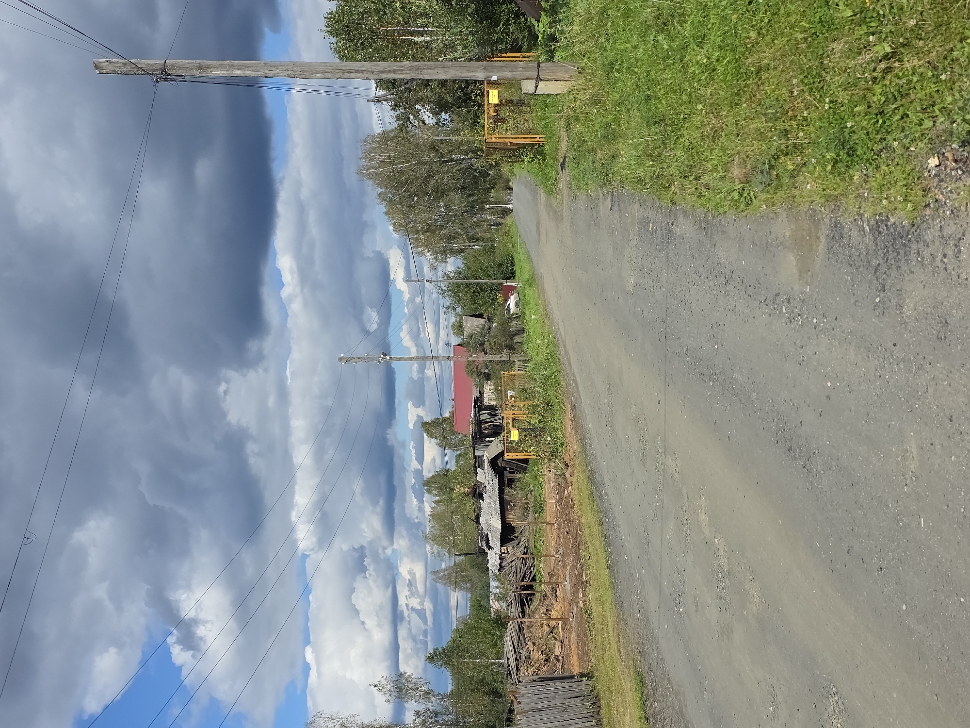 The image depicts a rural road in Magnitka, Russia, under a cloudy sky. The road is unpaved and appears to be a dirt and gravel path. On the left side of the road, there is a dilapidated wooden fence and a pile of lumber and debris, suggesting an abandoned or under-construction property. Beyond this, several rustic wooden houses with metal roofs are visible, some appearing to be in disrepair. Trees and foliage line the sides of the road and surround the houses. On the right side of the road, a wooden utility pole with power lines stretches across the frame. Further down the road, a yellow metal gate stands open, bordering a grassy area. The overall scene conveys a sense of rural isolation and a modest, possibly aging, community.