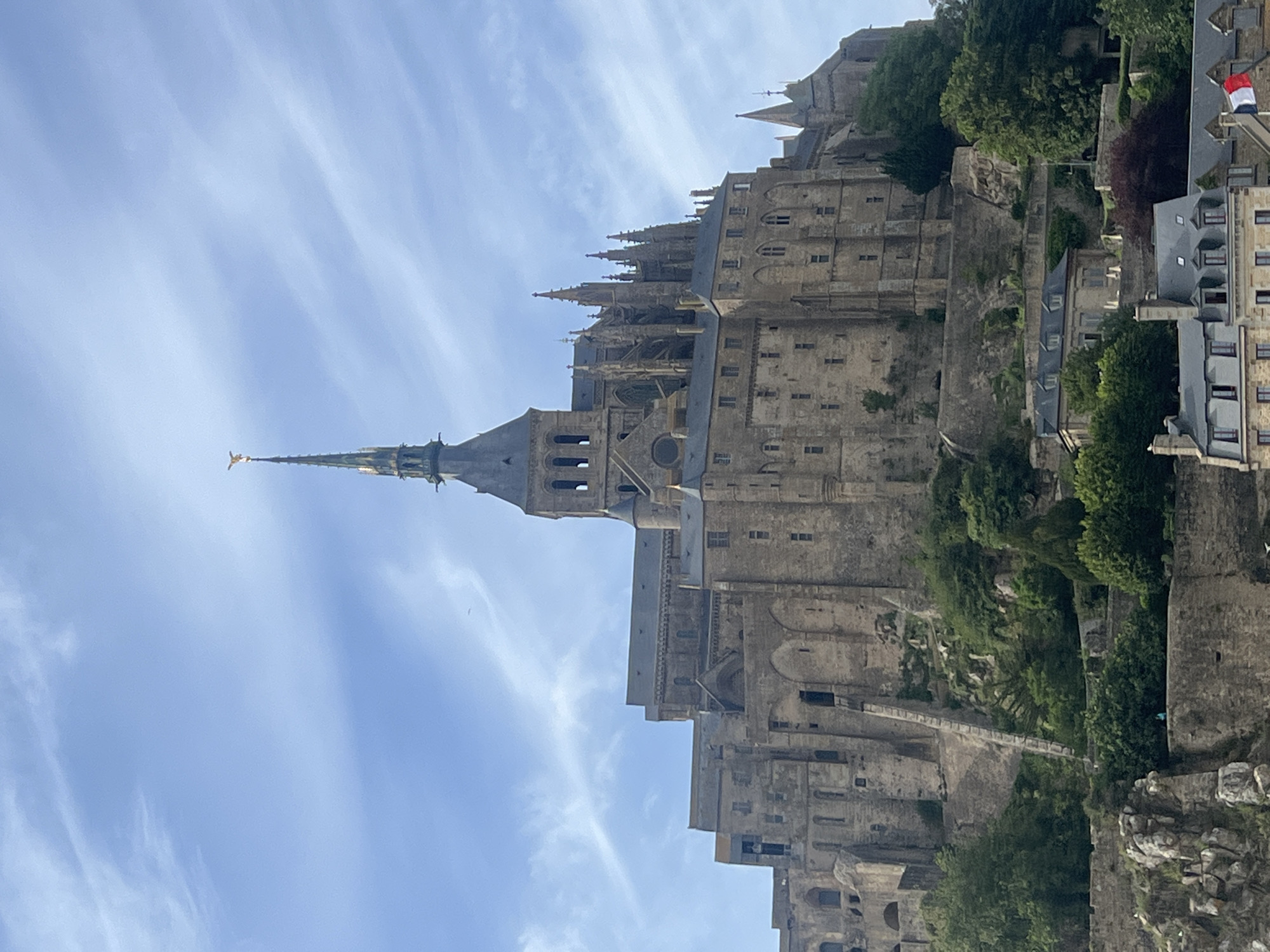 This image depicts the Mont Saint-Michel Abbey in Le Mont-Saint-Michel, France, under a clear sky with scattered wispy clouds. The massive medieval stone structure, primarily gray-brown, occupies the central and upper portions of the frame, built atop a natural rock formation covered in green foliage.
The abbey features a prominent central spire, topped with a golden statue, likely Archangel Michael. Gothic architectural elements are evident across the structure, including multiple smaller spires, pointed arches, various windows, and complex stone facades. Terraced sections of the abbey descend the rocky slope, revealing different architectural styles and building phases. A long, diagonal stone ramp or staircase is visible on the left side of the main structure, ascending the slope.
Below the main abbey, nestled among dense green trees and bushes, are several smaller, older stone buildings with dark gray roofs. One of these lower buildings prominently displays a small French flag. On the chimney of another lower building, a white bird is perched. In the upper left quadrant of the sky, a small, dark flying object is visible. The overall scene conveys a historical, fortified architectural complex integrated into a natural, elevated landscape.