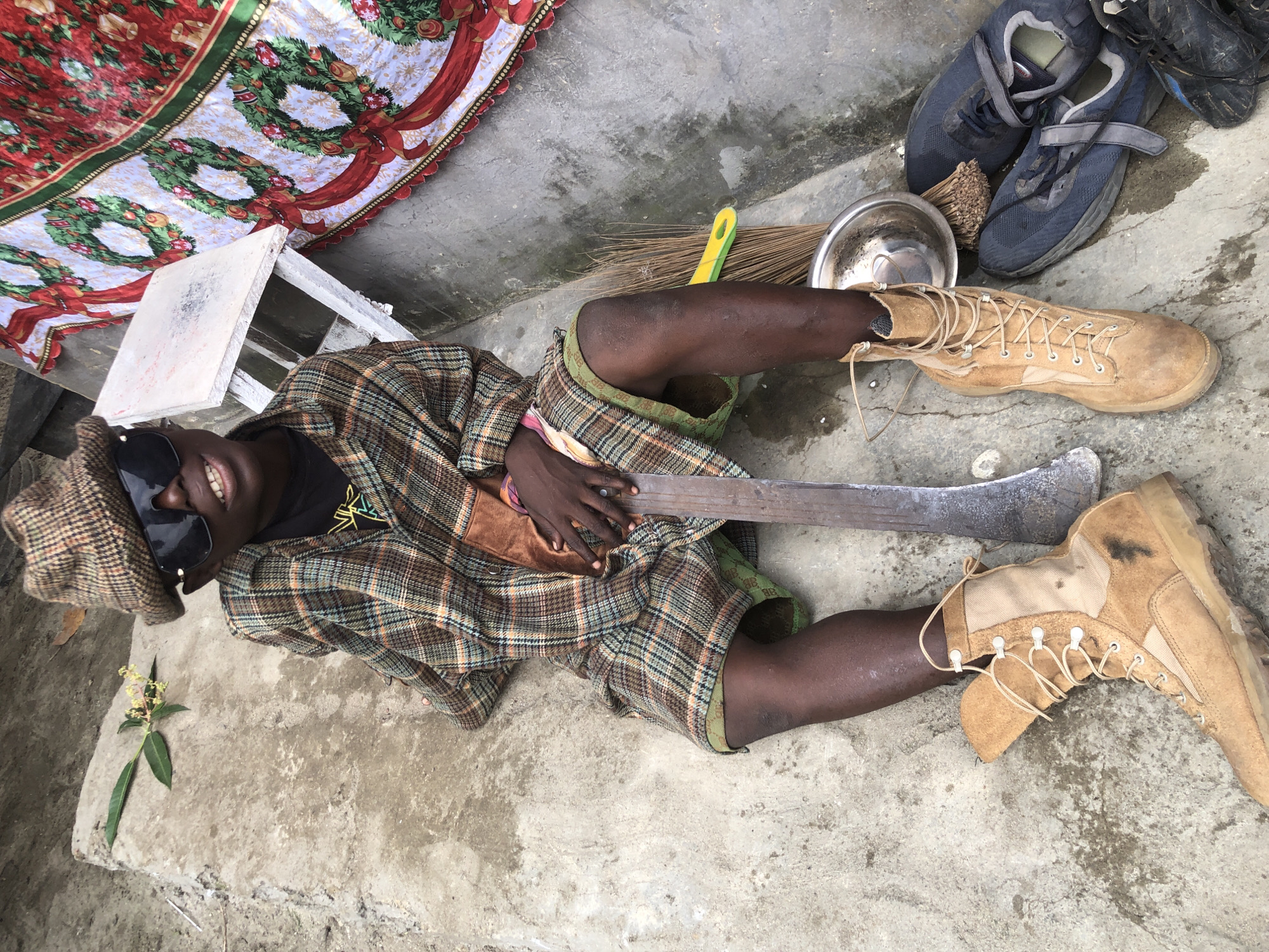 A dark-skinned individual, possibly male, wearing a plaid shirt, plaid hat, and sunglasses, is seated on a concrete surface. The individual is holding a large, flat metal tool, possibly a machete or a piece of farm equipment, which is resting on their leg. There is a broom and a metal bowl nearby. To the right, a decorative cloth with images of wreaths and bells is draped over a white chair. The environment appears to be outdoors, with the concrete ground showing some signs of wear and dampness. The background includes a wall and some greenery. The scene is set in Ado, Nigeria.