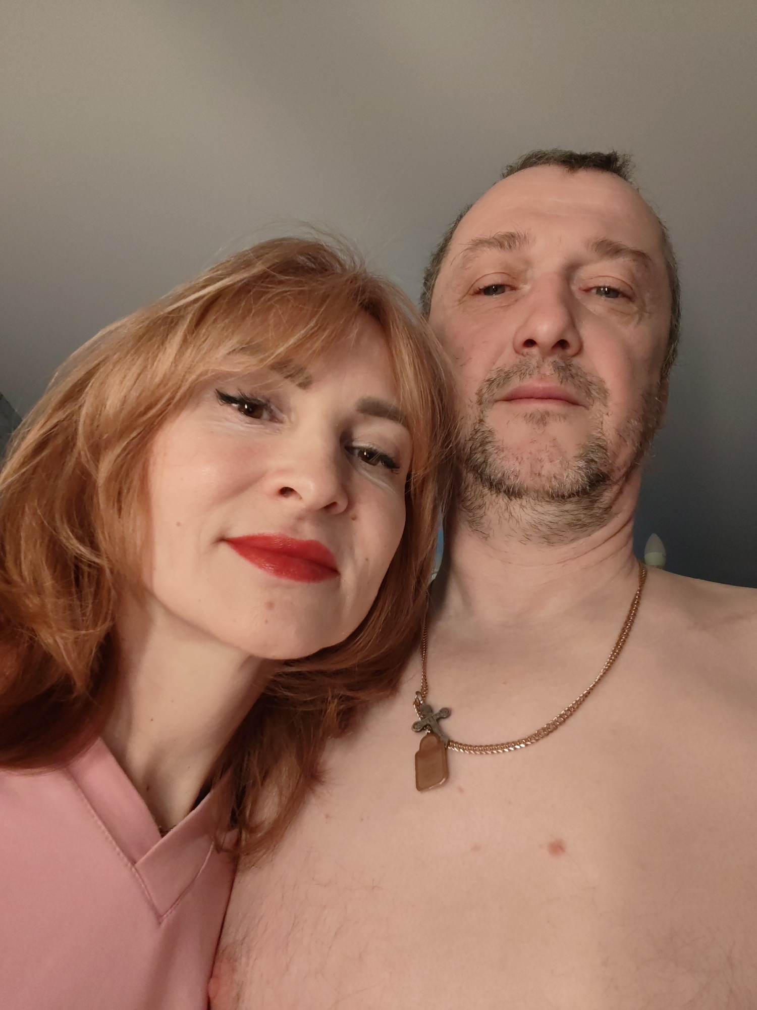 A photograph depicts a man and a woman posing closely together, taken at an indoor location, likely in an apartment setting based on the background. The woman is on the left, with shoulder-length red hair and wearing bright red lipstick. She has fair skin and is wearing a pink top. The man is on the right, with short grey hair, facial stubble, and is shirtless, wearing a gold chain necklace with a cross pendant. The background is a plain, light-colored wall. There is no indication of the location being in Chaikovsky, Russia.