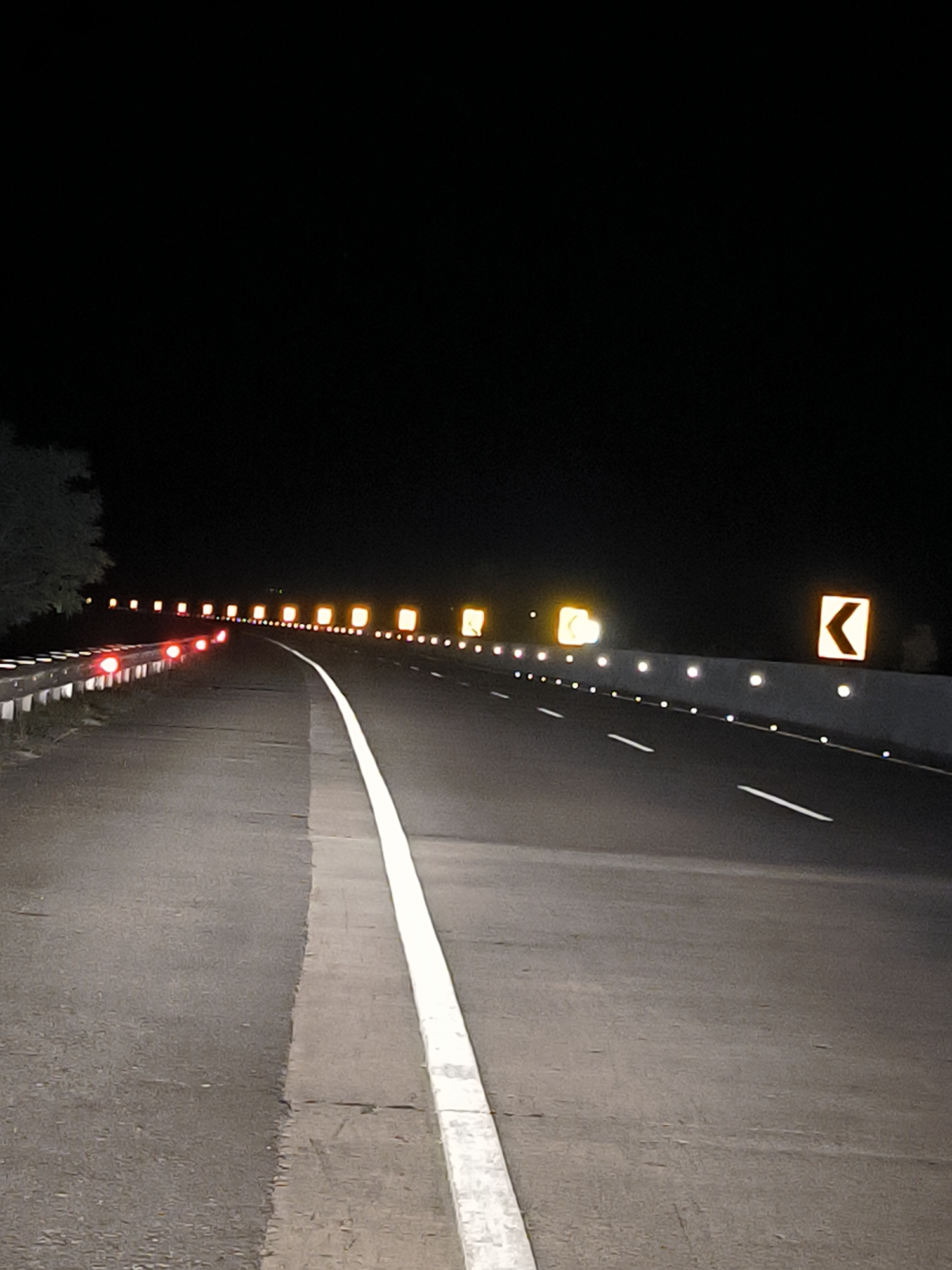 An eye-level, full shot captures a highway at night with a white line dividing two lanes of asphalt. The lane on the left is bordered by a metal guardrail with several red reflectors. Along the right side of the highway, a series of elevated, yellow rectangular signs with black arrows indicate a curve to the left, interspersed with small, white reflective markers. The sky above is completely black. The scene appears to be located on a highway in the vicinity of Keureuweungkrueng, Indonesia.