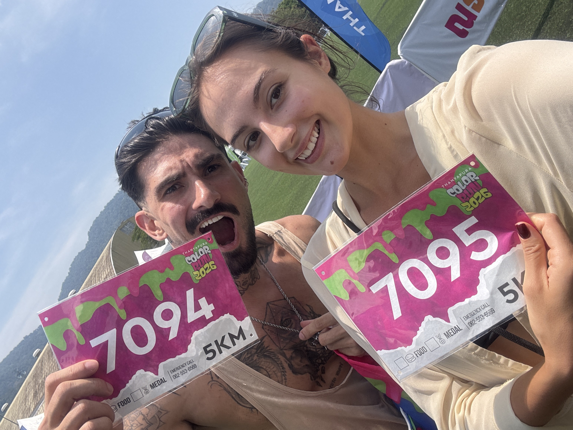 A close-up, angled shot shows a couple holding up race bibs at the "Thanyapura Color Run 2026" 5K event. The man on the left is fair-skinned with dark hair and a beard; he's wearing a tan tank top and has visible tattoos on his chest and arms. His mouth is open in an excited expression. The woman on the right is also fair-skinned with dark hair, wearing sunglasses on her head and a light-colored top. She's smiling at the camera. They are holding up pink and white race bibs; his bib number is "7094", hers is "7095". In the background, there's a grassy field, tents, and trees. The location is Thanyapura in Thep Krasatti, Thailand.